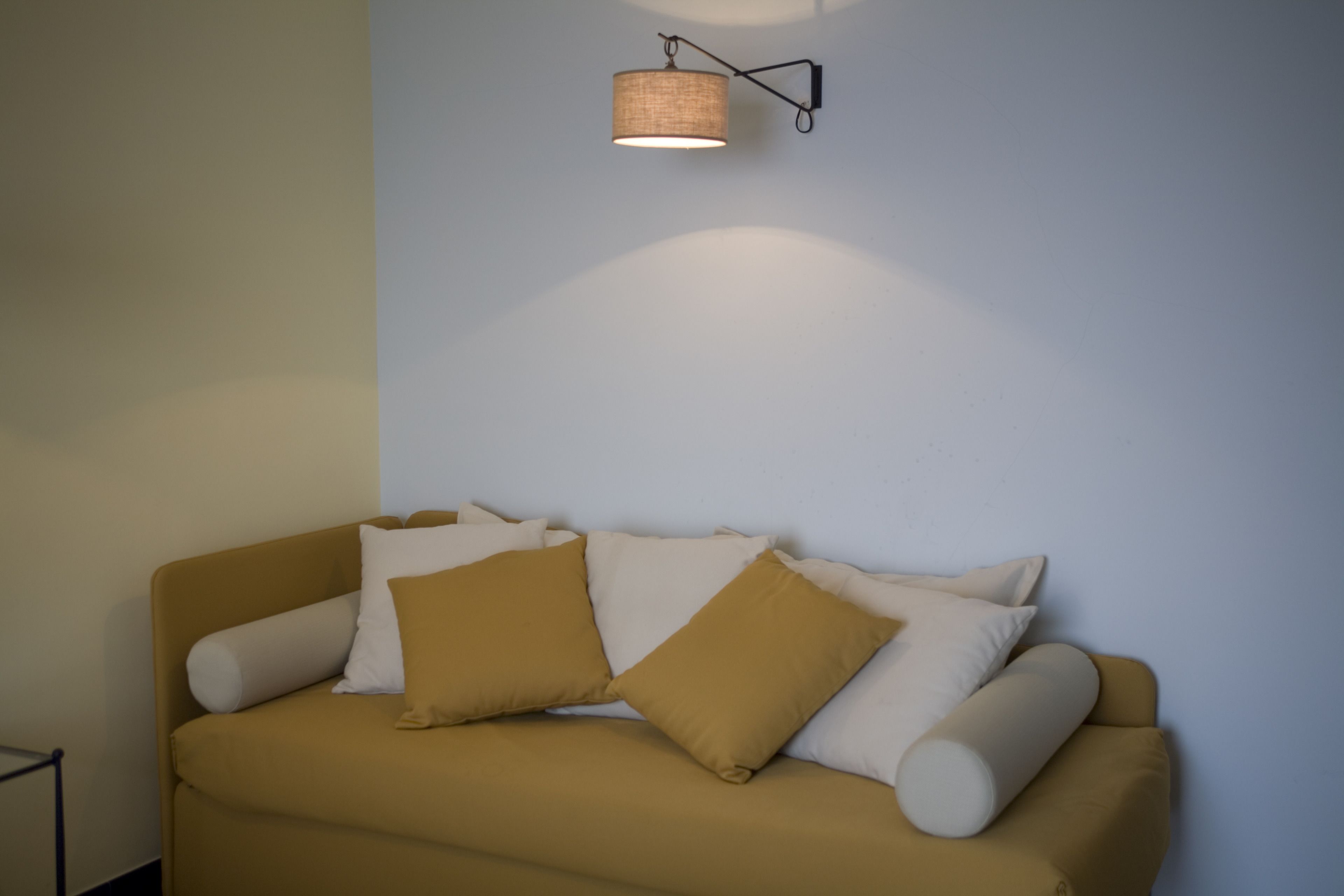 Un sofá beige con varios cojines frente a una pared lisa, una mesita a la izquierda y una lámpara de pared arriba.