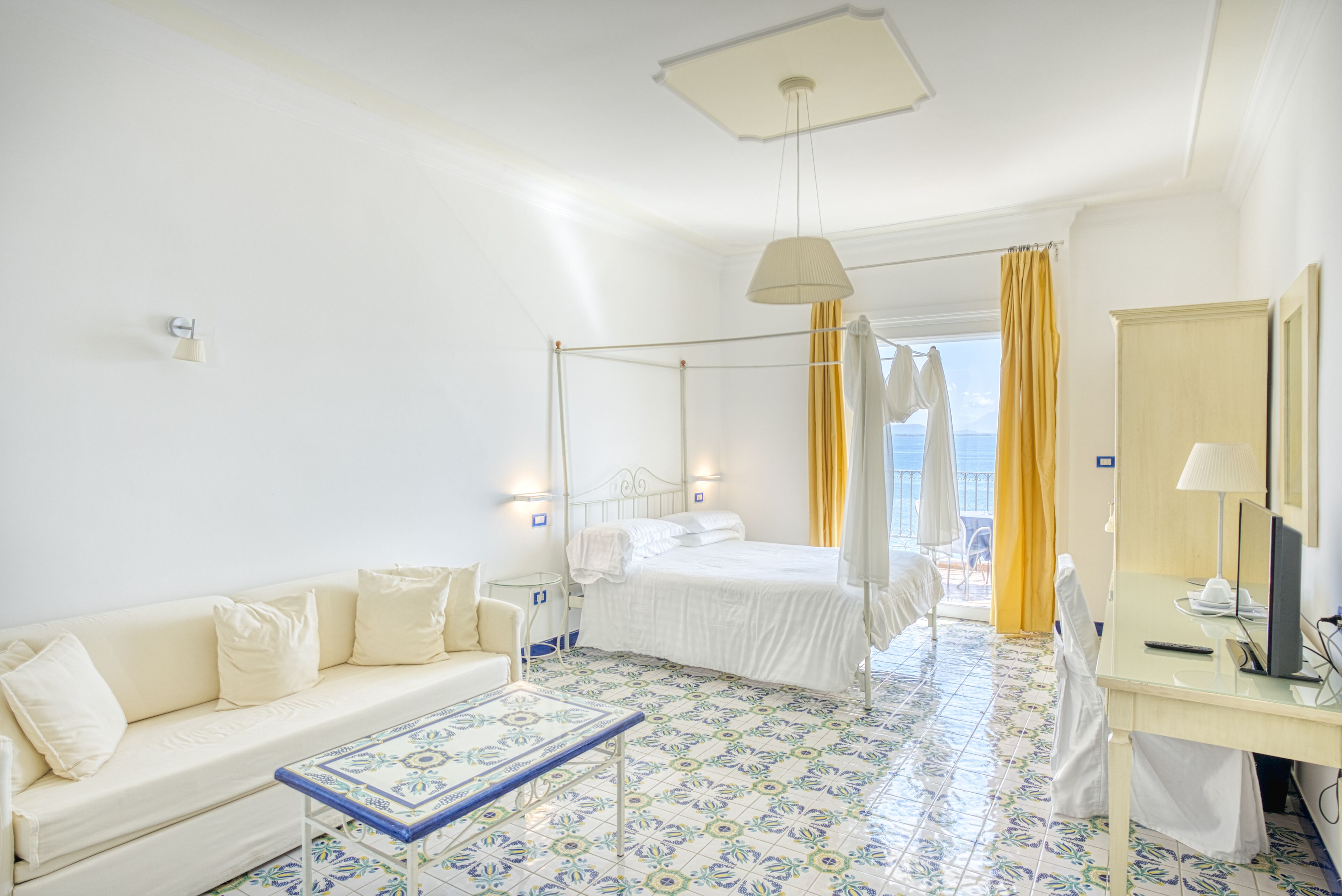 A hotel room with a bed, a sofa, a desk with a TV, and a view of the sea through the window.