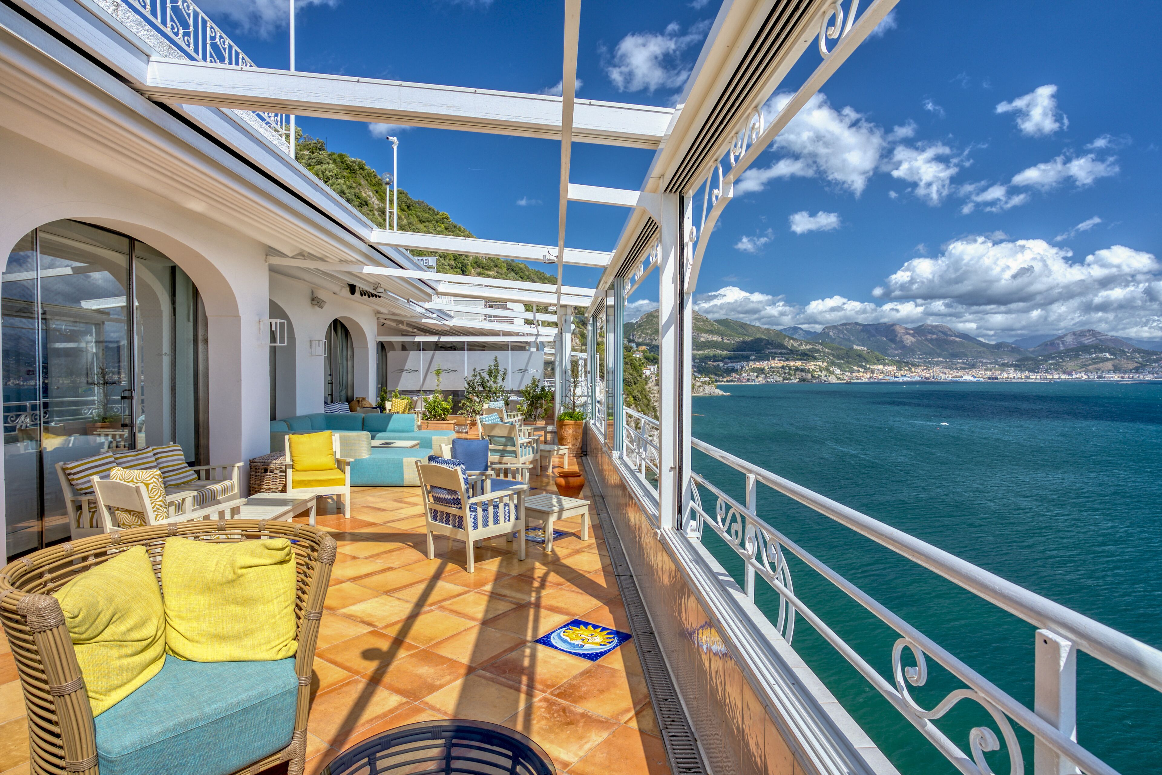 A sunlit deck with outdoor seating, a view of the sea, and a cityscape in the distance.