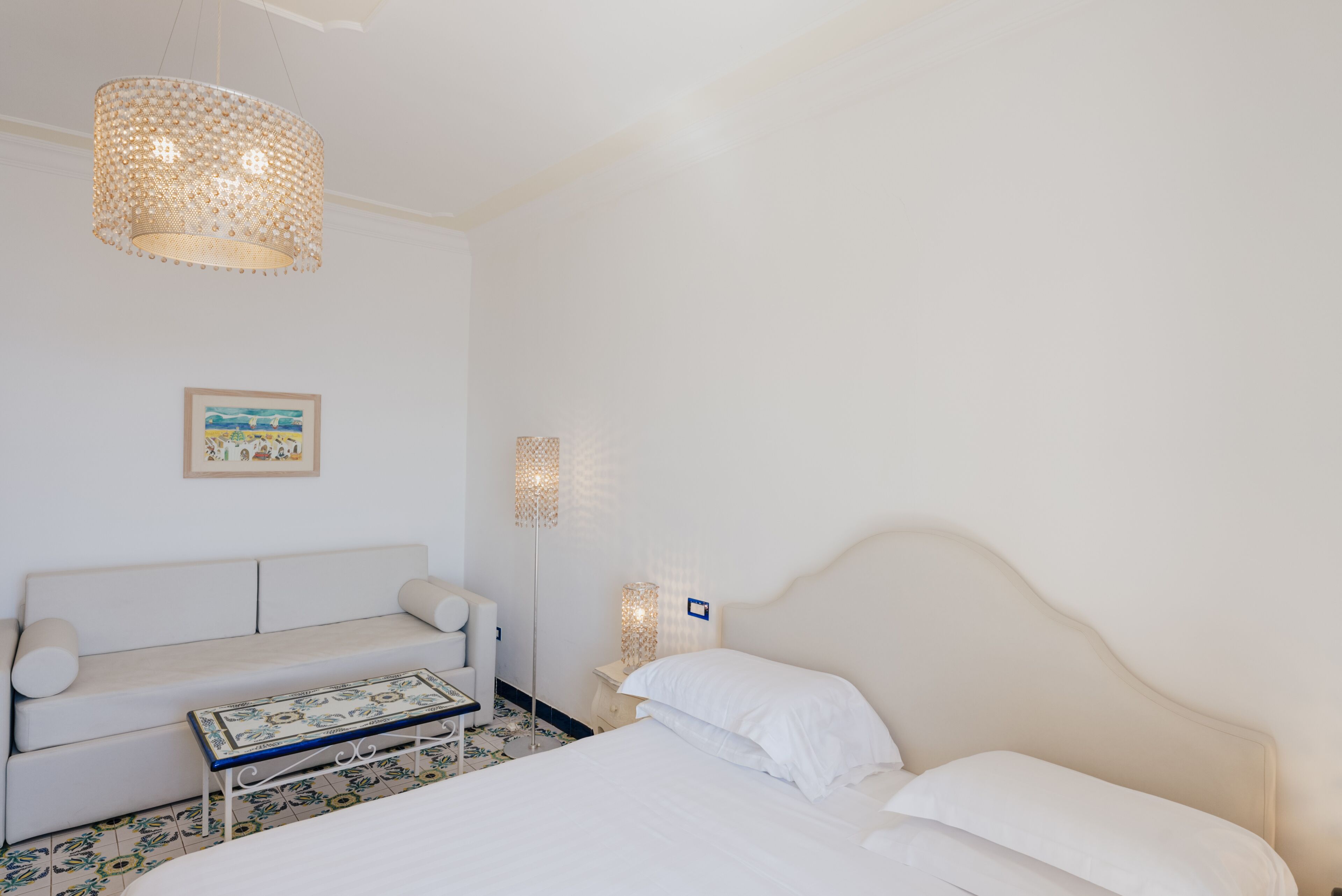 A hotel room with a bed, a sofa, a small table, a floor lamp, and a wall-mounted light.