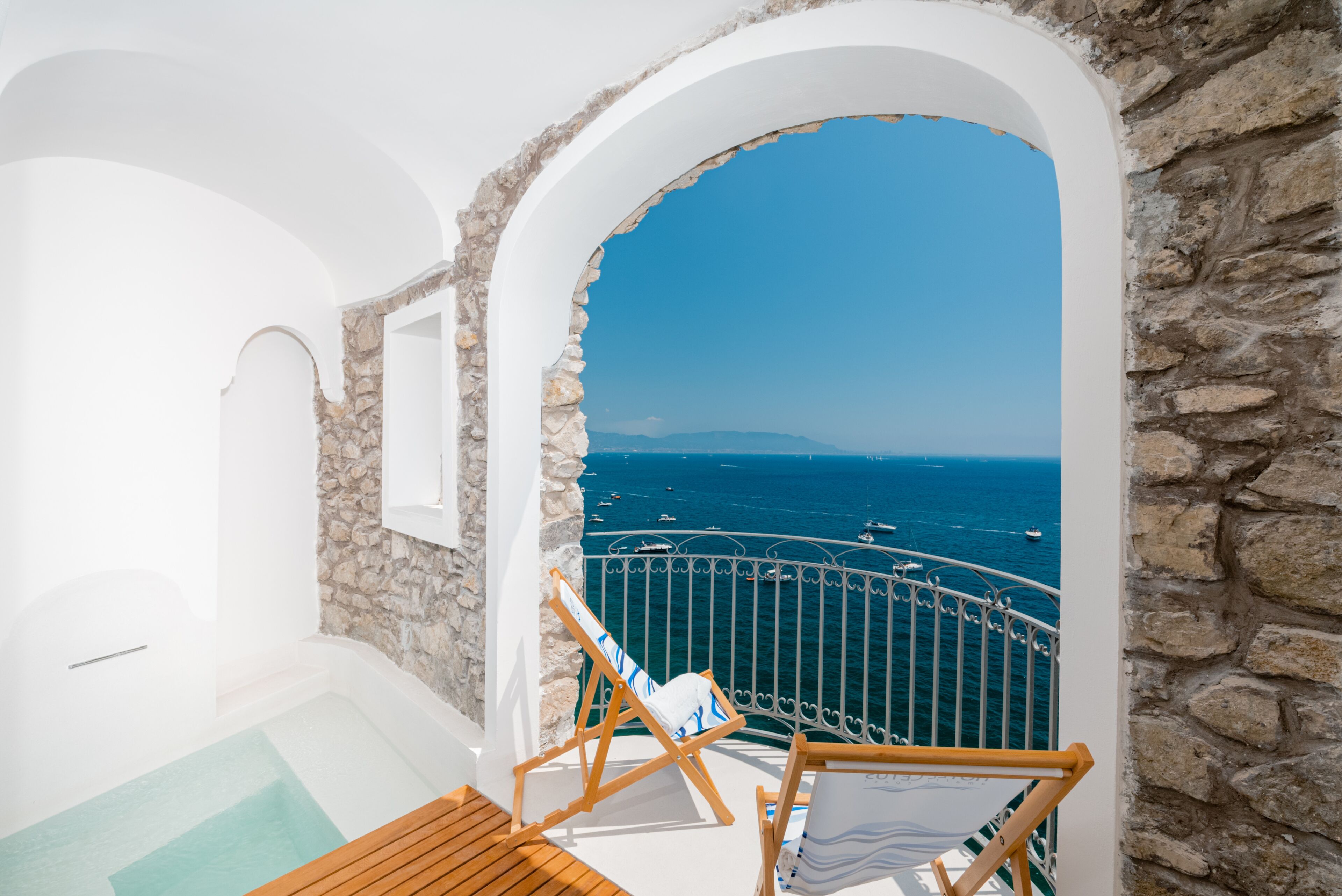 A balcony with a stone wall, a wooden deck, and a view of the sea with boats.