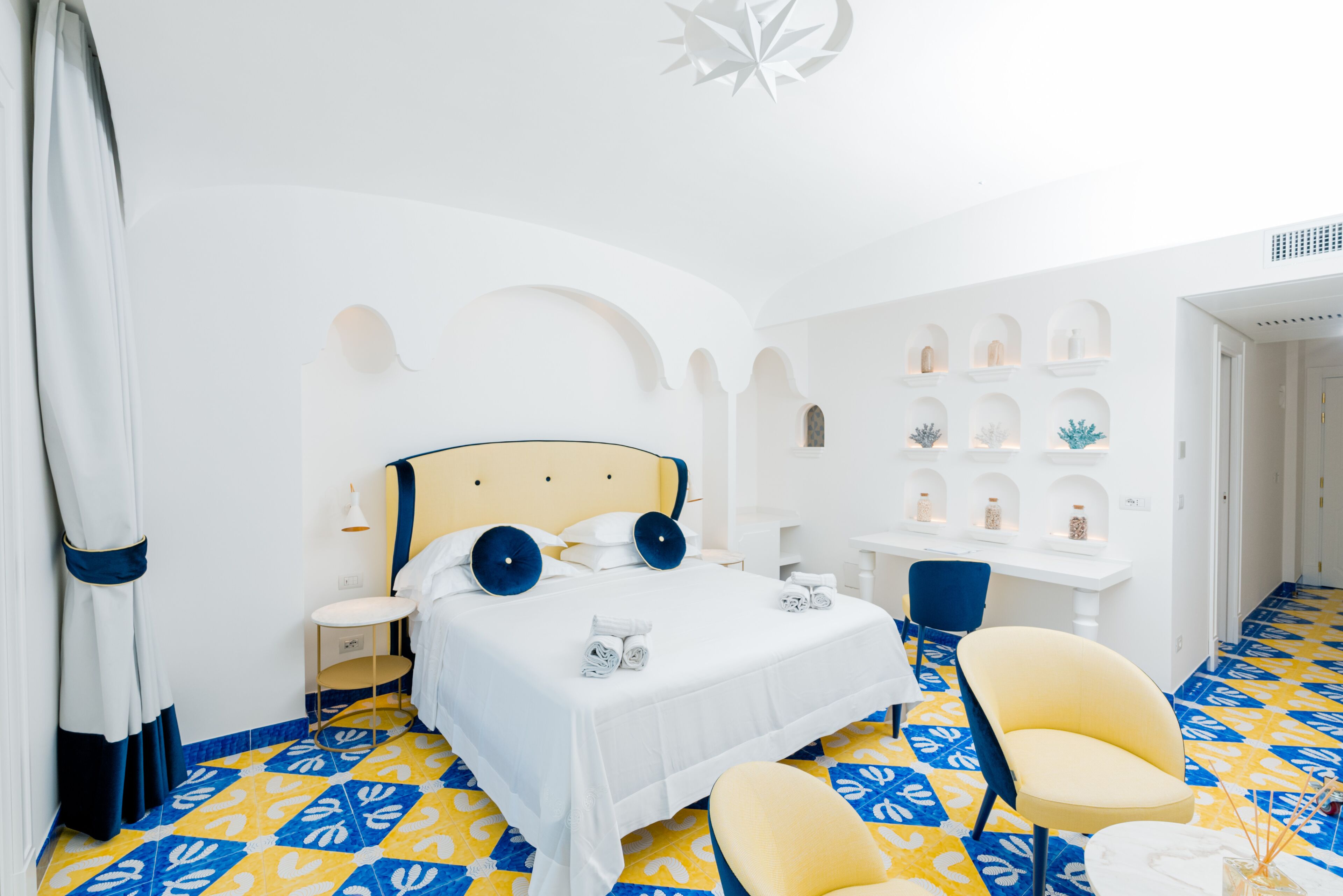A hotel room with a bed, a chair, a desk, and shelves with decorative items.