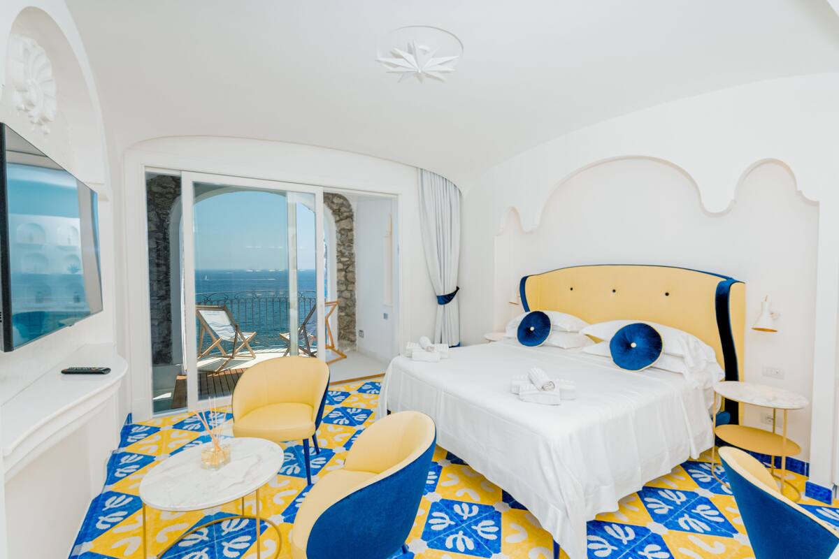 A hotel room with a bed, a chair, a small table, and a balcony with a view of the sea.
