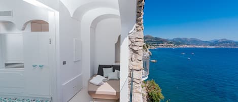 A balcony with a wooden floor, white walls, and a stone pillar. A bed with pillows and a towel is visible. The balcony overlooks a body of water with boats and distant land.
