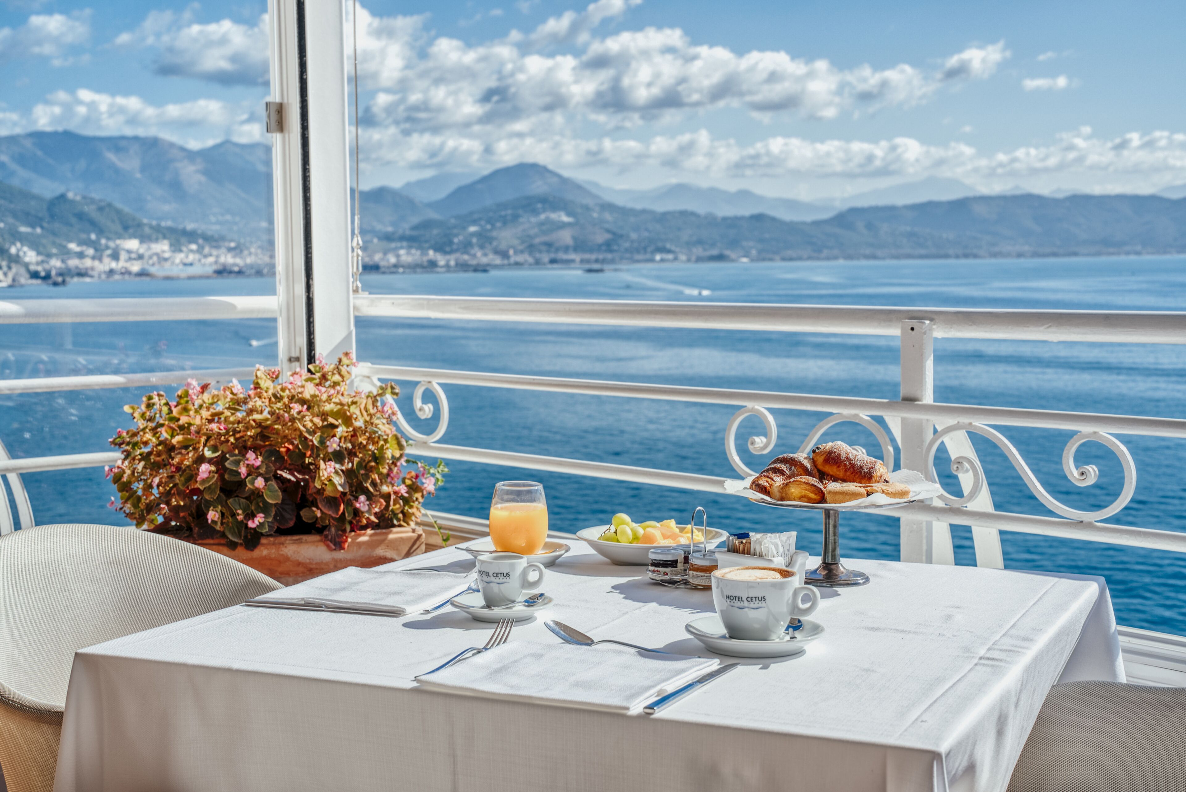 A table set with breakfast items, a flower arrangement, and a scenic view of the sea and mountains.