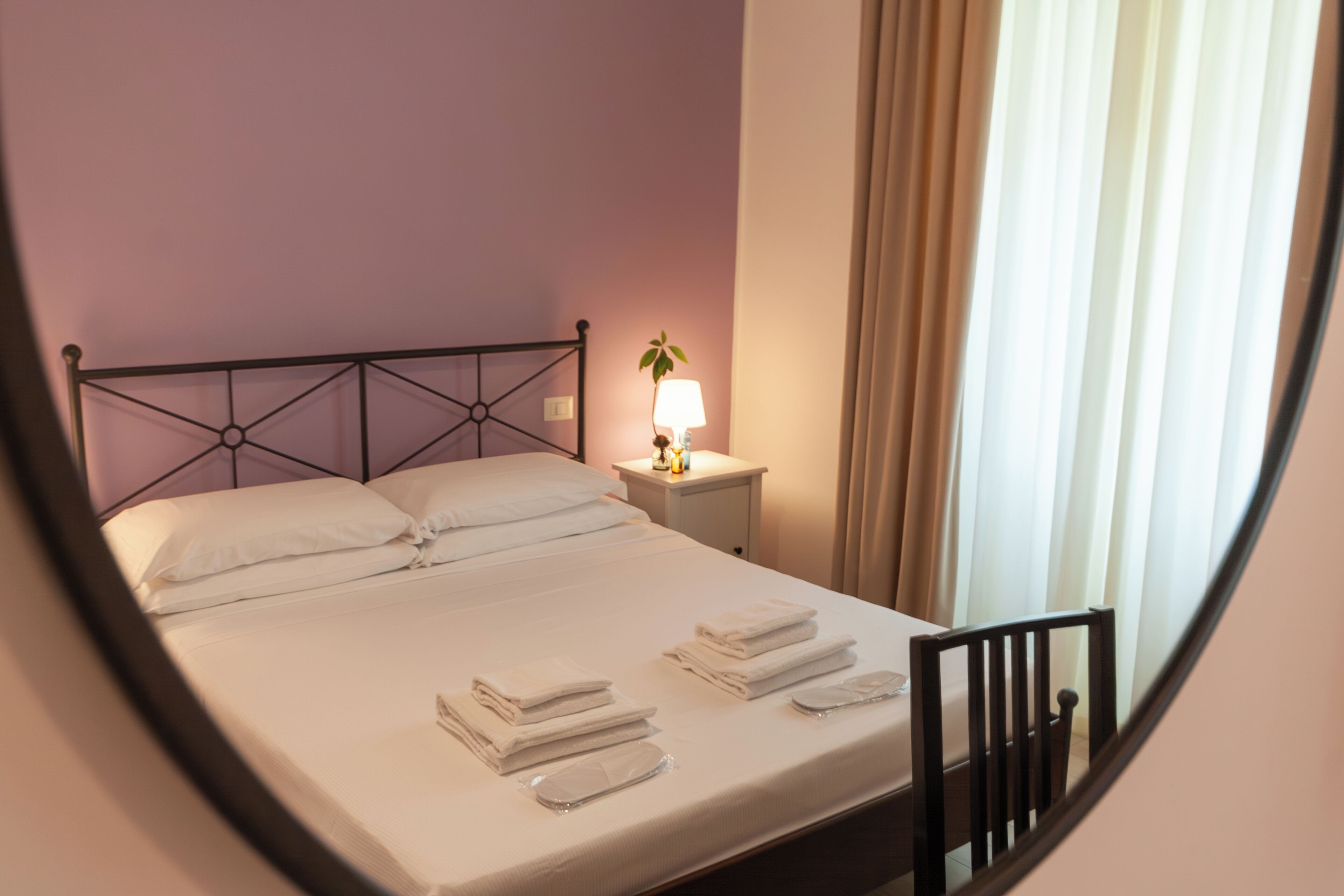 A neatly made bed with white linens and towels, a bedside table with a lamp, and a window with curtains.