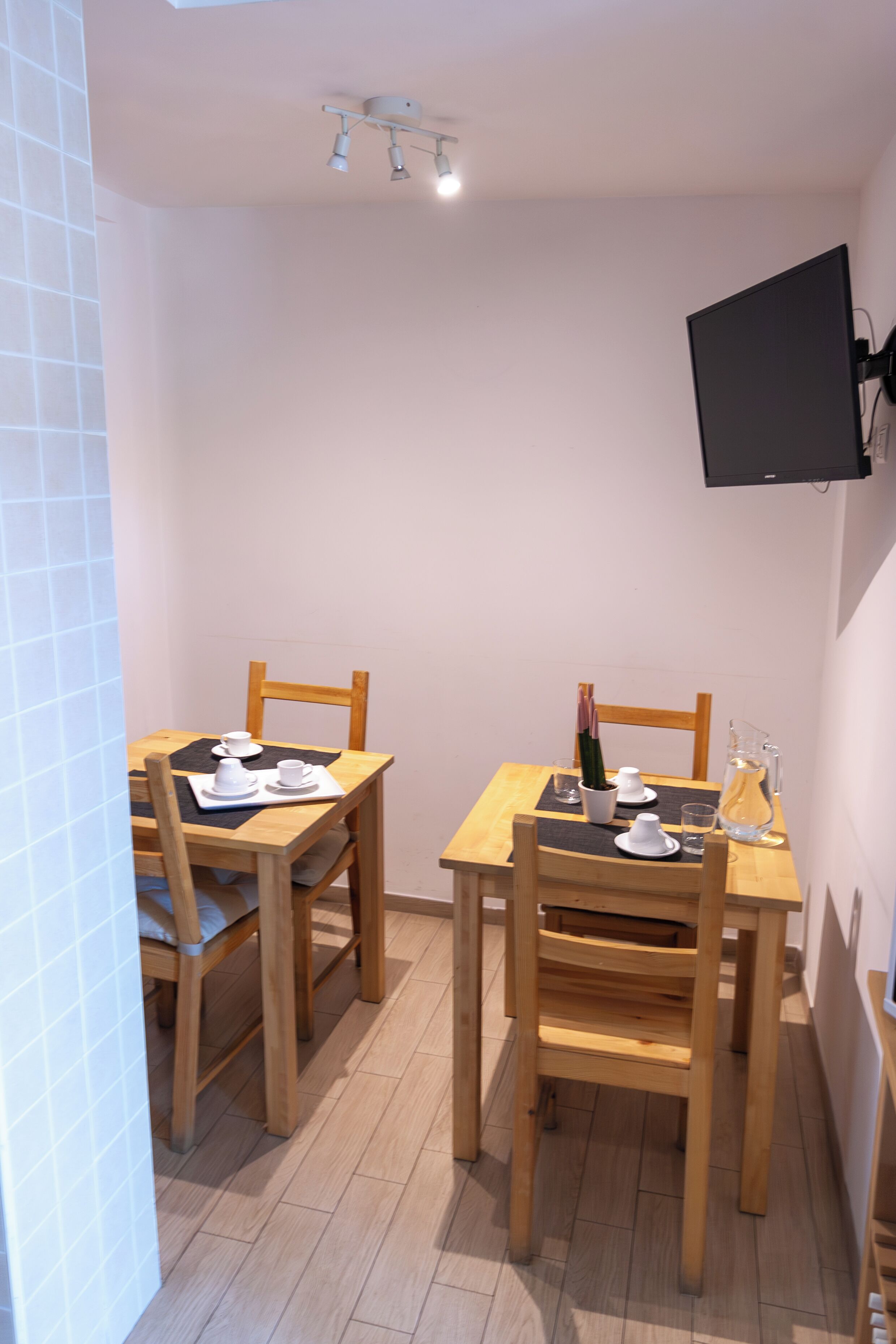 A small dining area with two wooden tables and chairs, a flat-screen TV mounted on the wall, and a tiled floor.