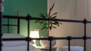Una cama con ropa blanca, una lámpara de noche, una planta en maceta y una mesita de noche con toallas dobladas.