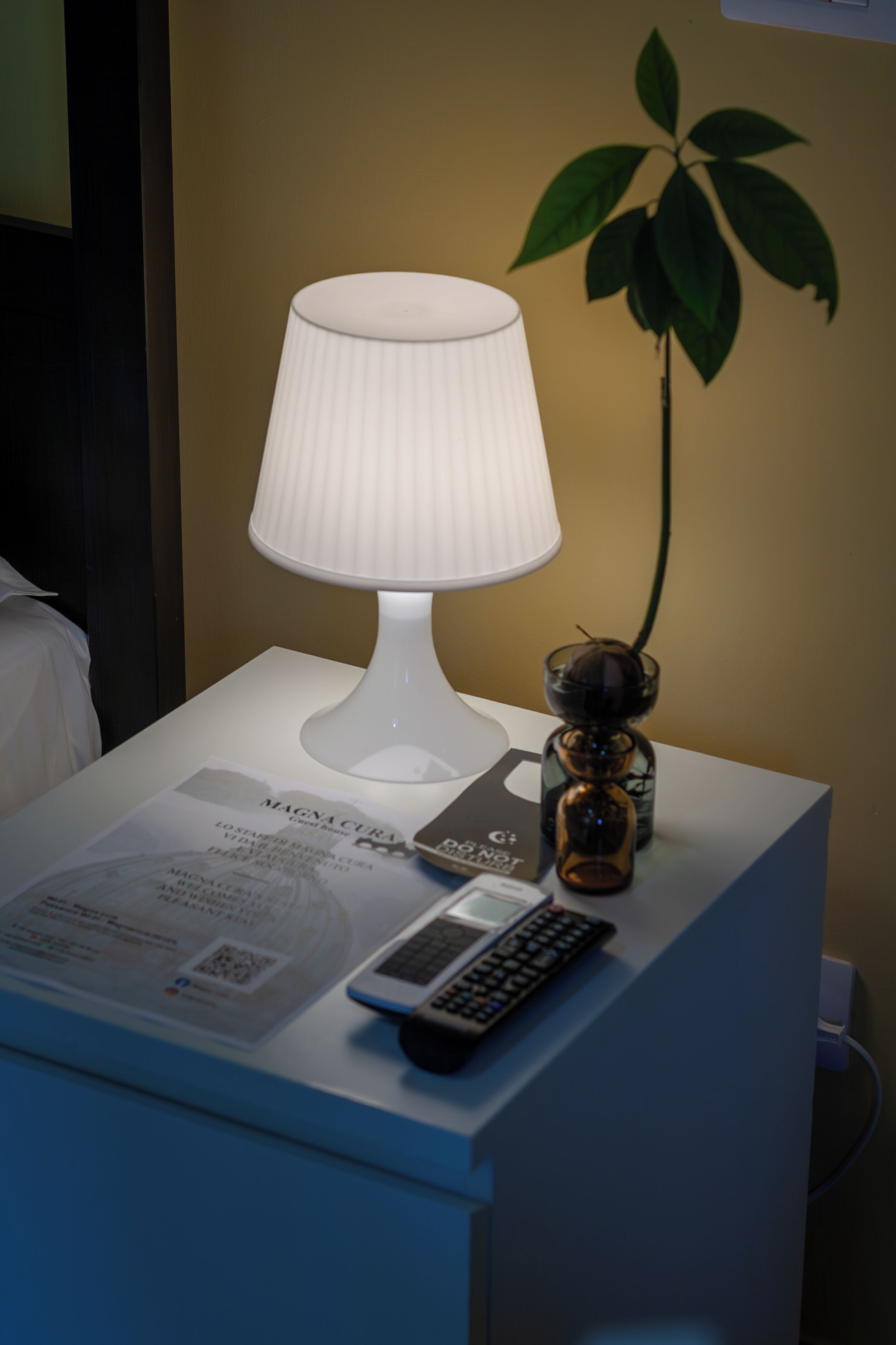 A bedside table with a white lamp, a small vase with a plant, a remote control, and a magazine.