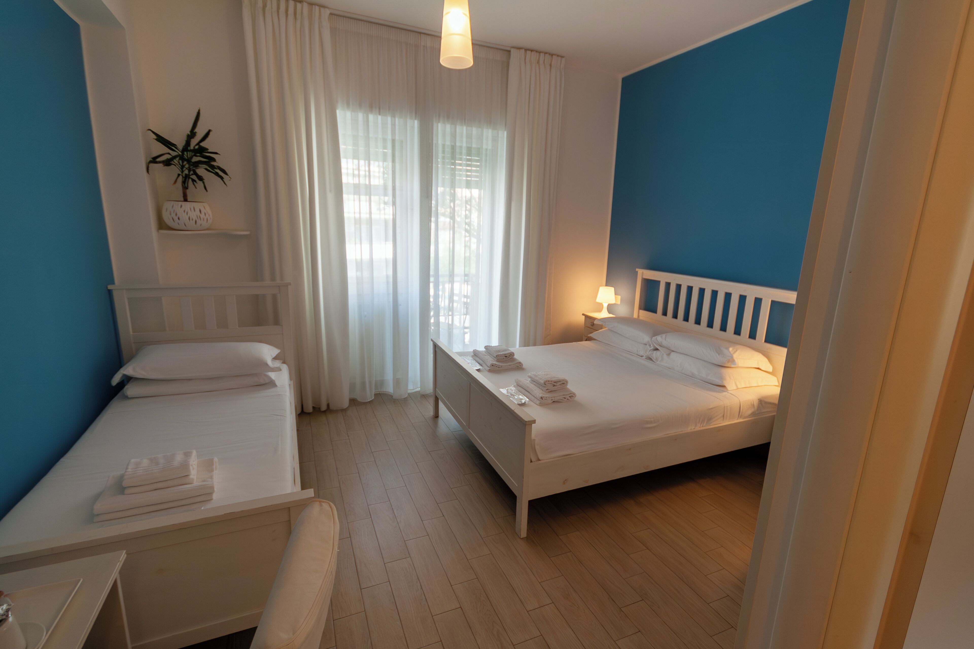 Habitació d'hotel amb dos llits, una taula de treball, una cadira, una planta en test i una finestra amb cortines.