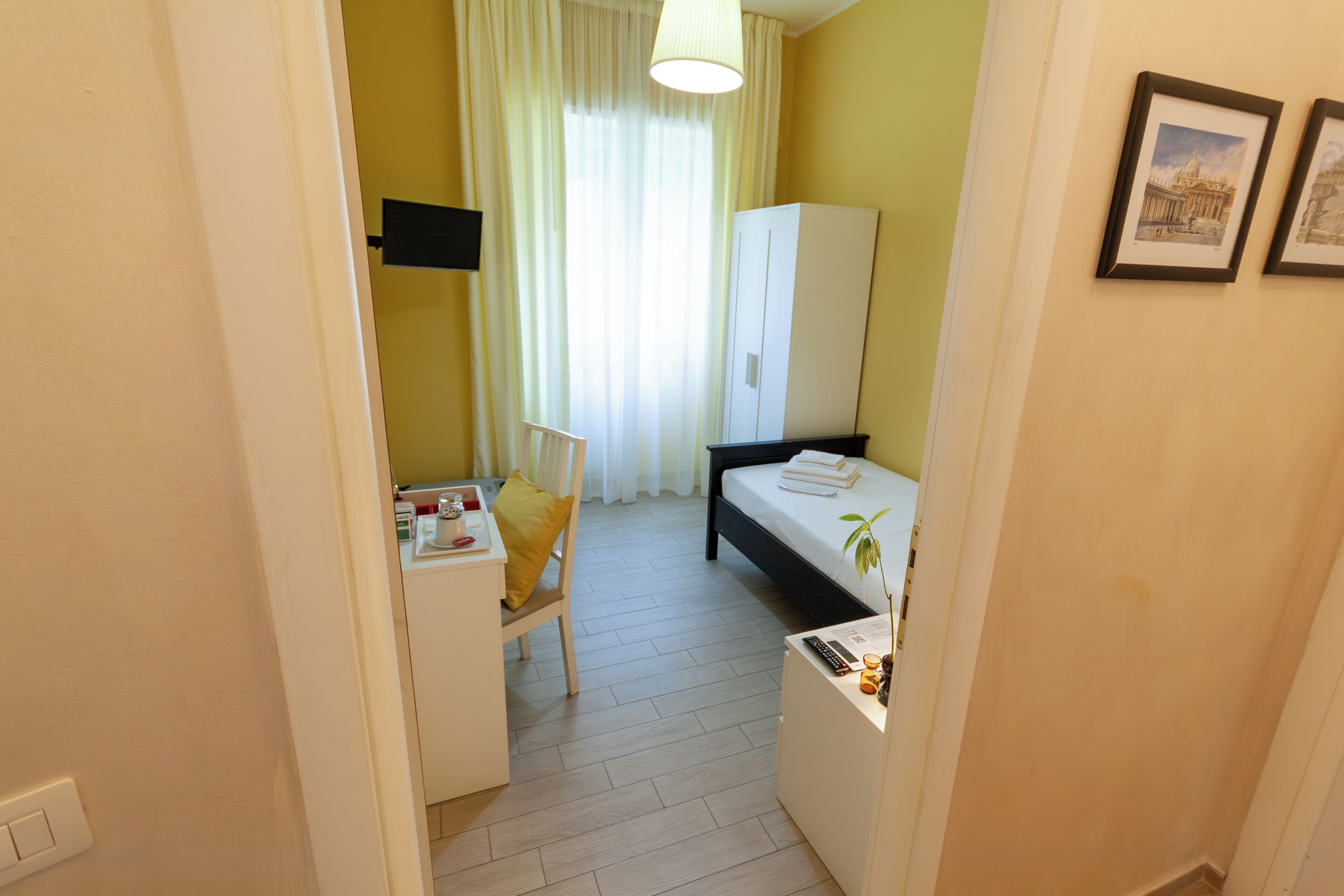 A compact hotel room with a bed, a desk, a chair, a wardrobe, and a small table with a plant.