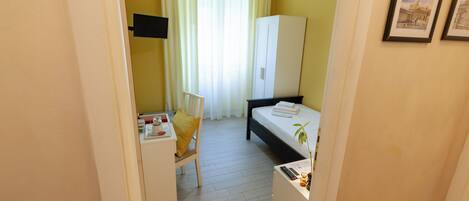 A compact hotel room with a bed, a desk, a chair, a wardrobe, and a small table with a plant.