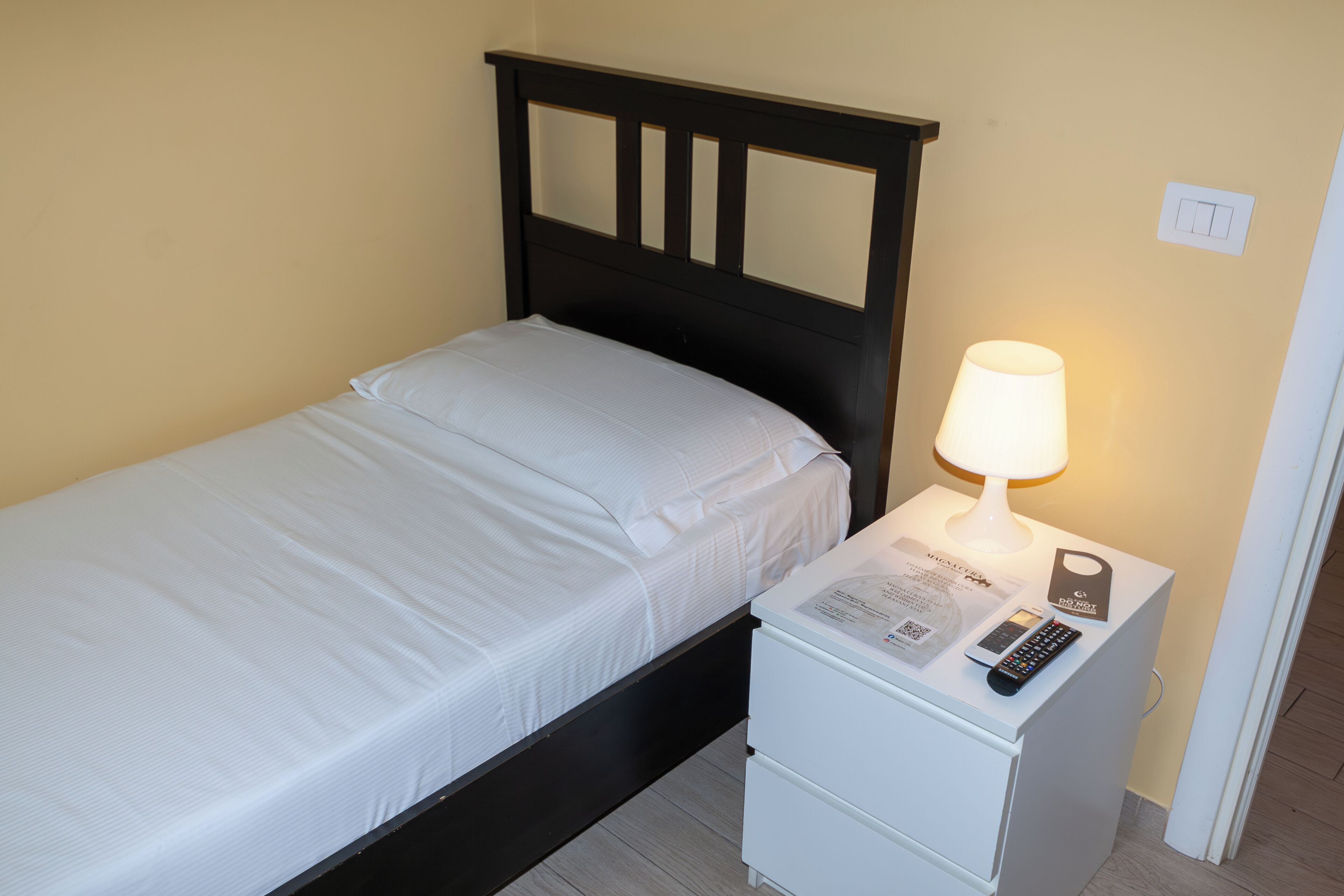 Un lit simple avec une tête de lit en bois, une table de chevet avec une lampe, une télécommande et une petite carte.