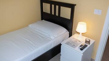 Una cama individual con cabecera de madera, una mesita de noche con lámpara, un control remoto y una pequeña tarjeta.