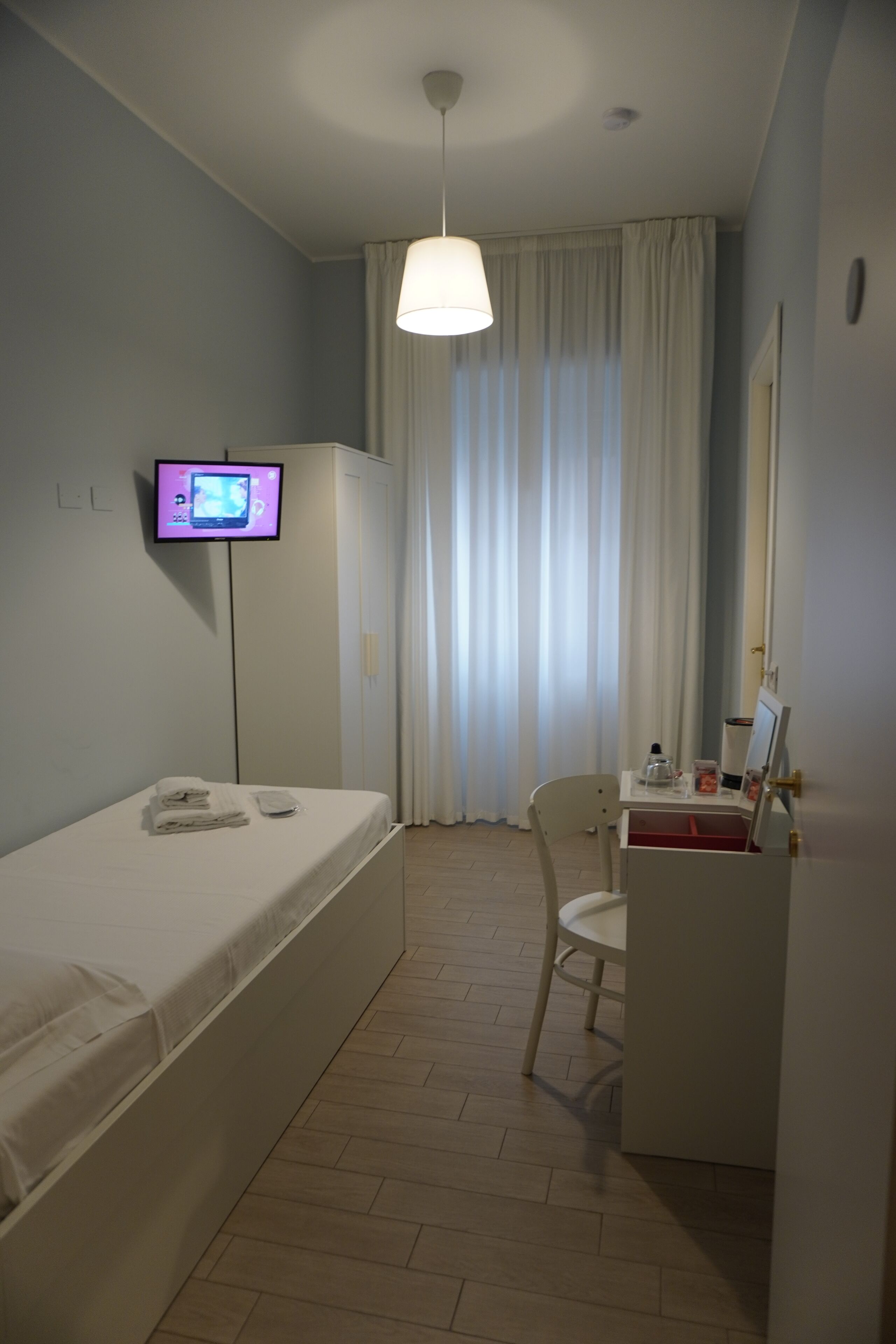 Une petite pièce, bien rangée, avec un lit, un bureau et une chaise, un téléviseur et une fenêtre avec des rideaux.