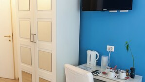 Una habitación con un escritorio blanco, una silla blanca, un televisor de pantalla plana montado en la pared, un armario blanco y suelo de madera.