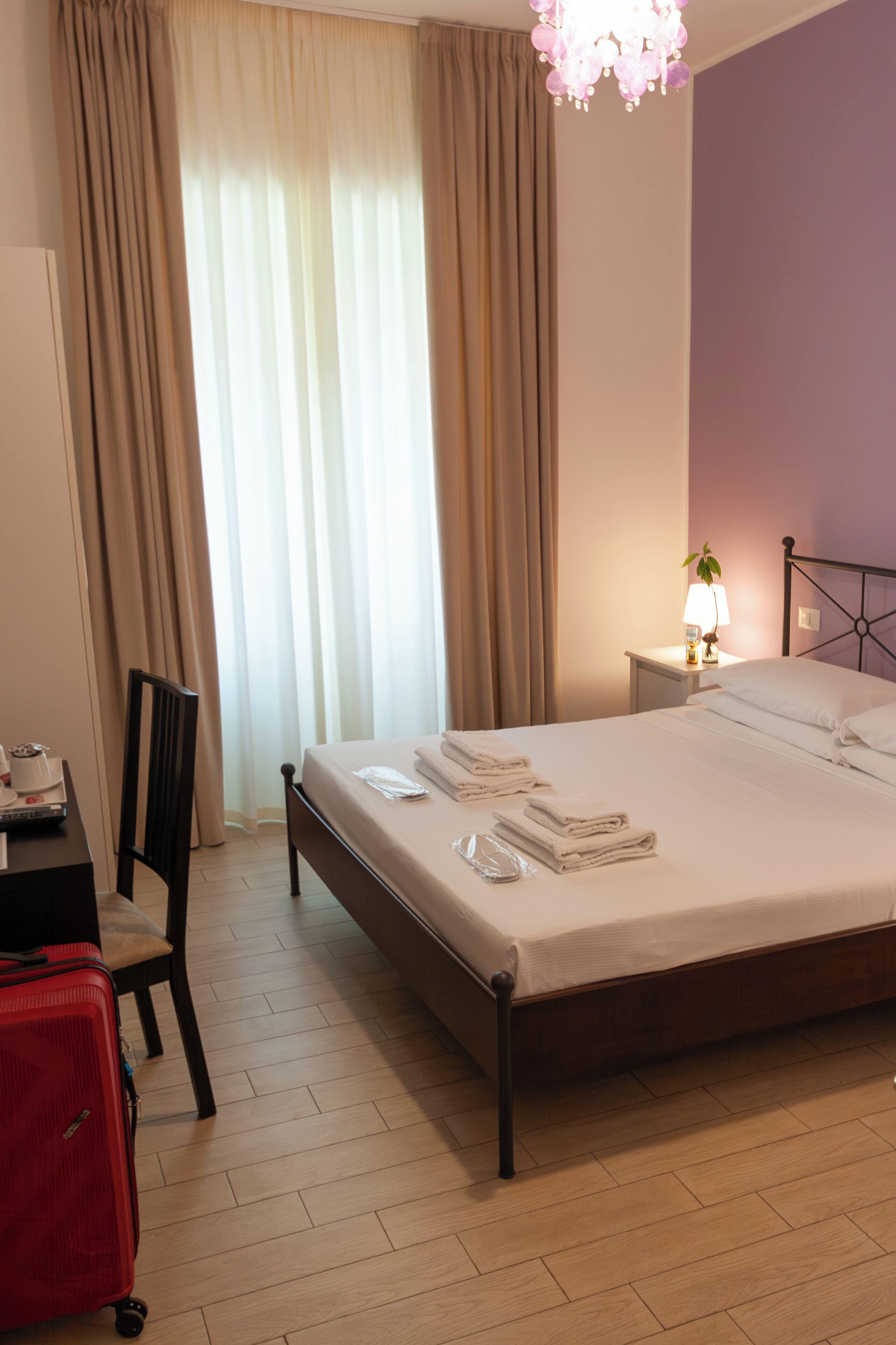 A hotel room with a bed, a desk with a chair, a red suitcase, a nightstand with a lamp, and a window with curtains.