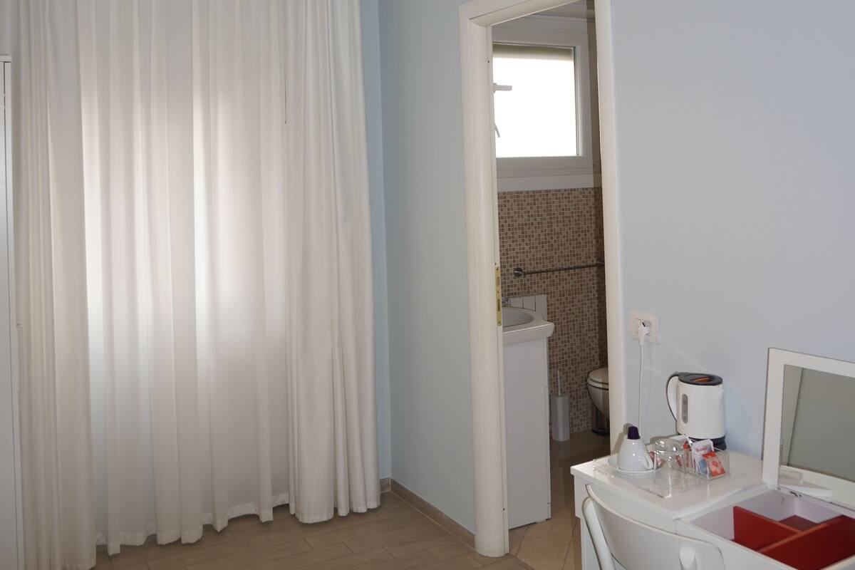 A hotel room with a bed, a desk with a chair, a white lamp, and a window with curtains.