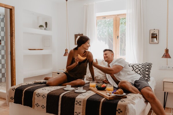 Una pareja sentada en una cama con alimentos para el desayuno, una ventana con cortinas y una pared blanca con estantes.