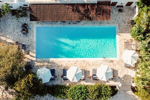 Vista aérea de una piscina con agua cristalina azul, rodeada de pavimento de piedra y sombrillas blancas.