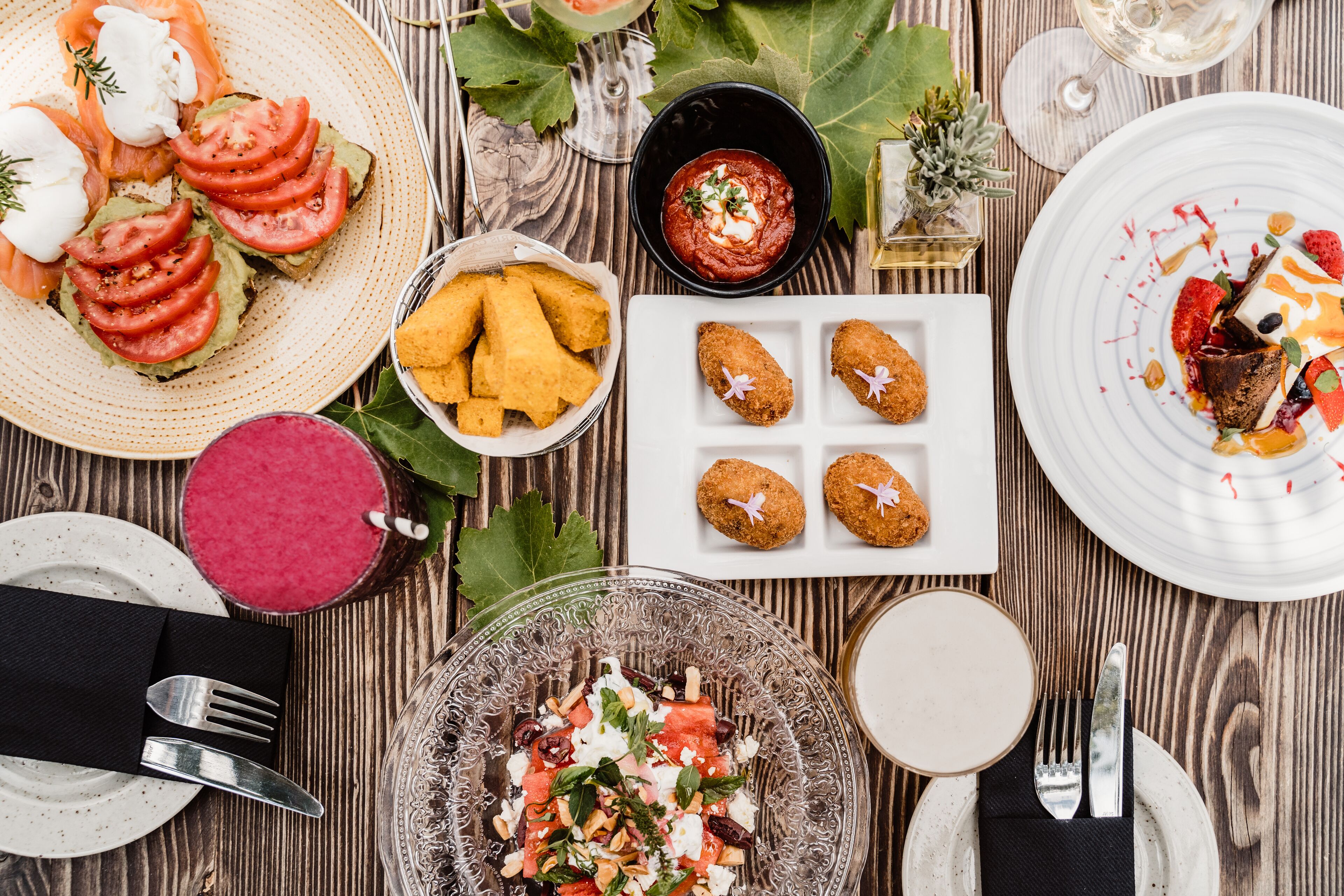 Una mesa de madera con diversos platos, entre ellos, una tostada de aguacate, un tazón de sopa y un plato de postre.