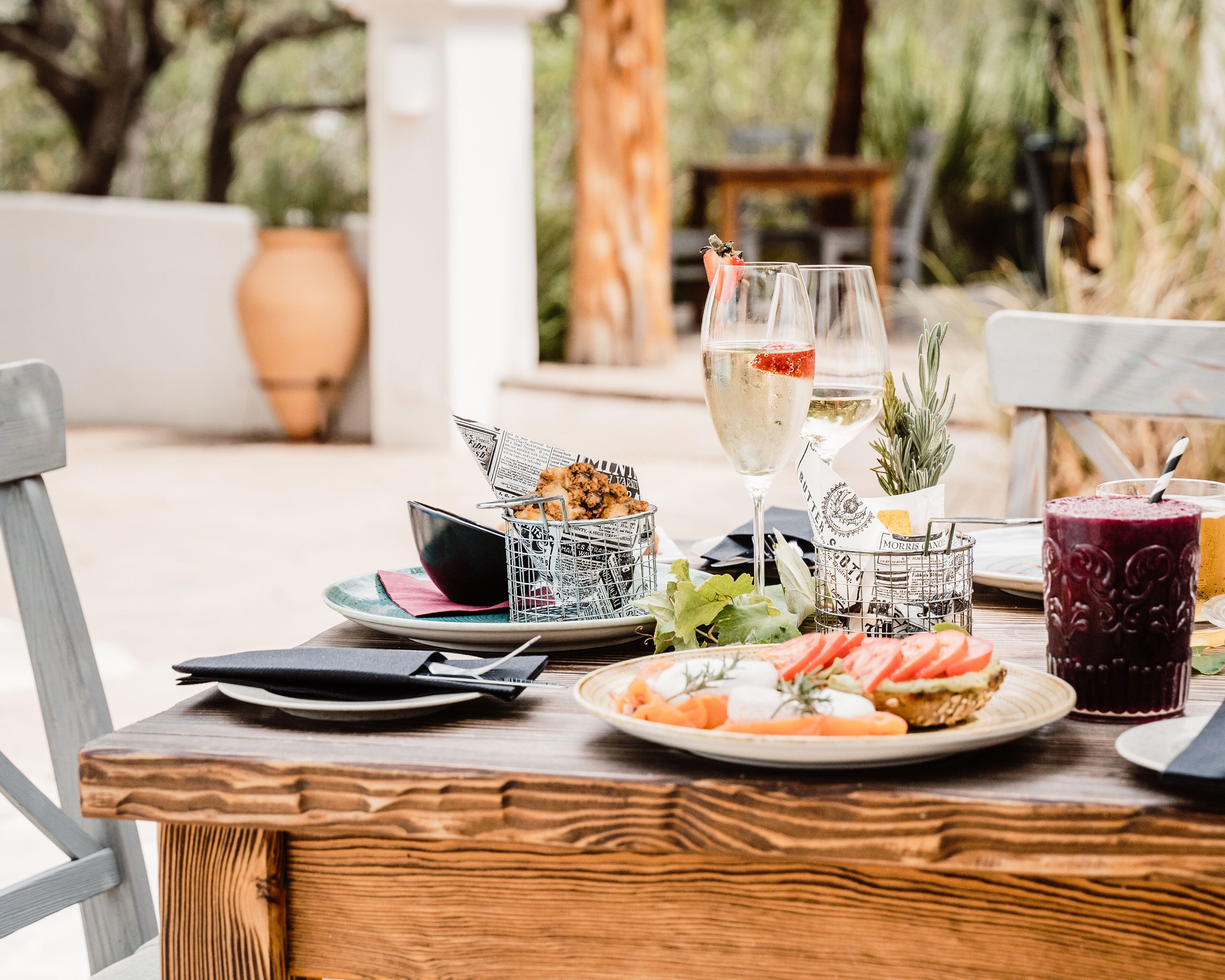 A wooden table set with a plate of food, two glasses of wine, a cocktail, and a menu.