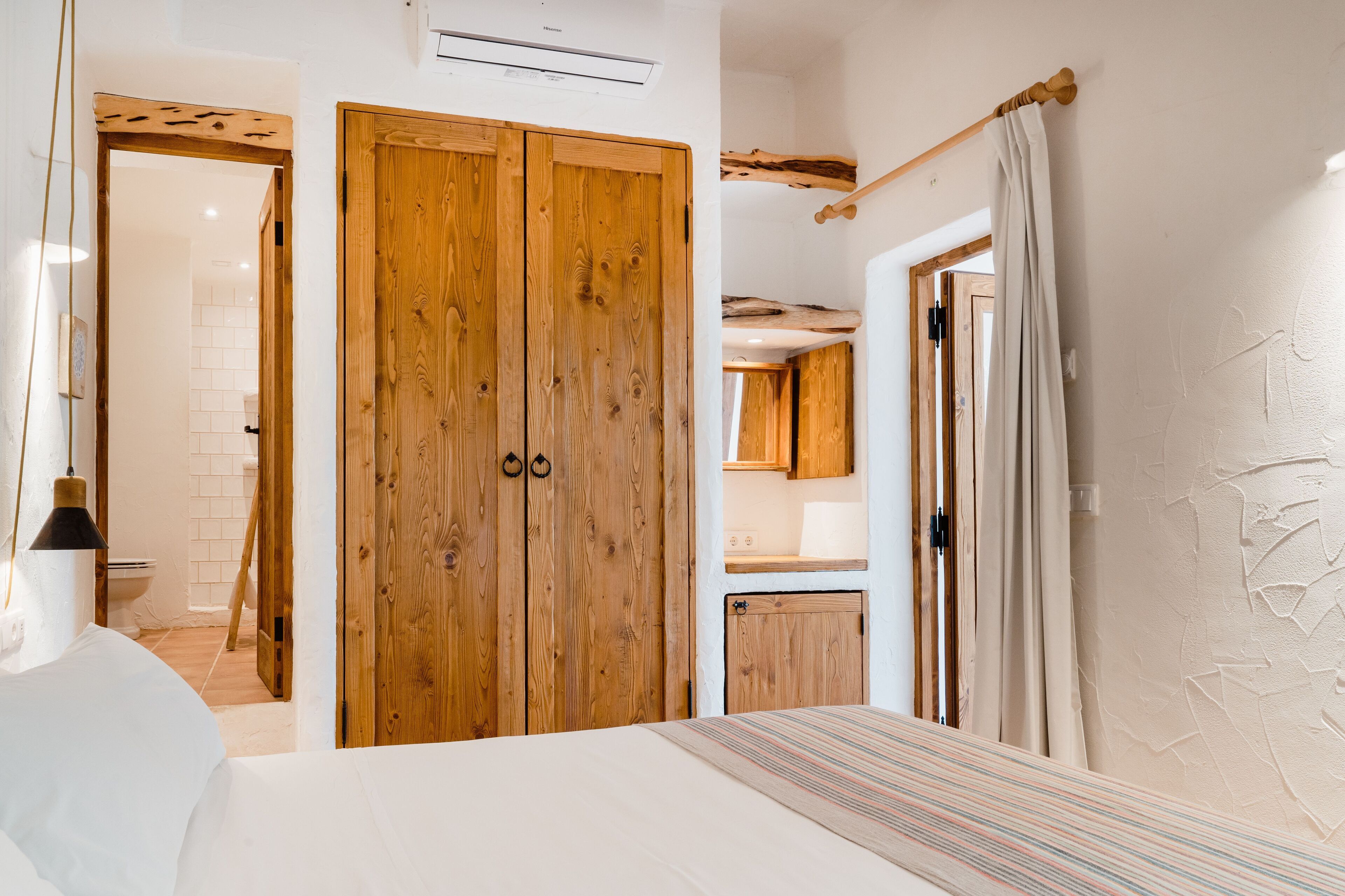 Une chambre à coucher avec une grande armoire en bois, un lit recouvert d’une couverture à rayures, et une vue sur la salle de bain et la cuisine.