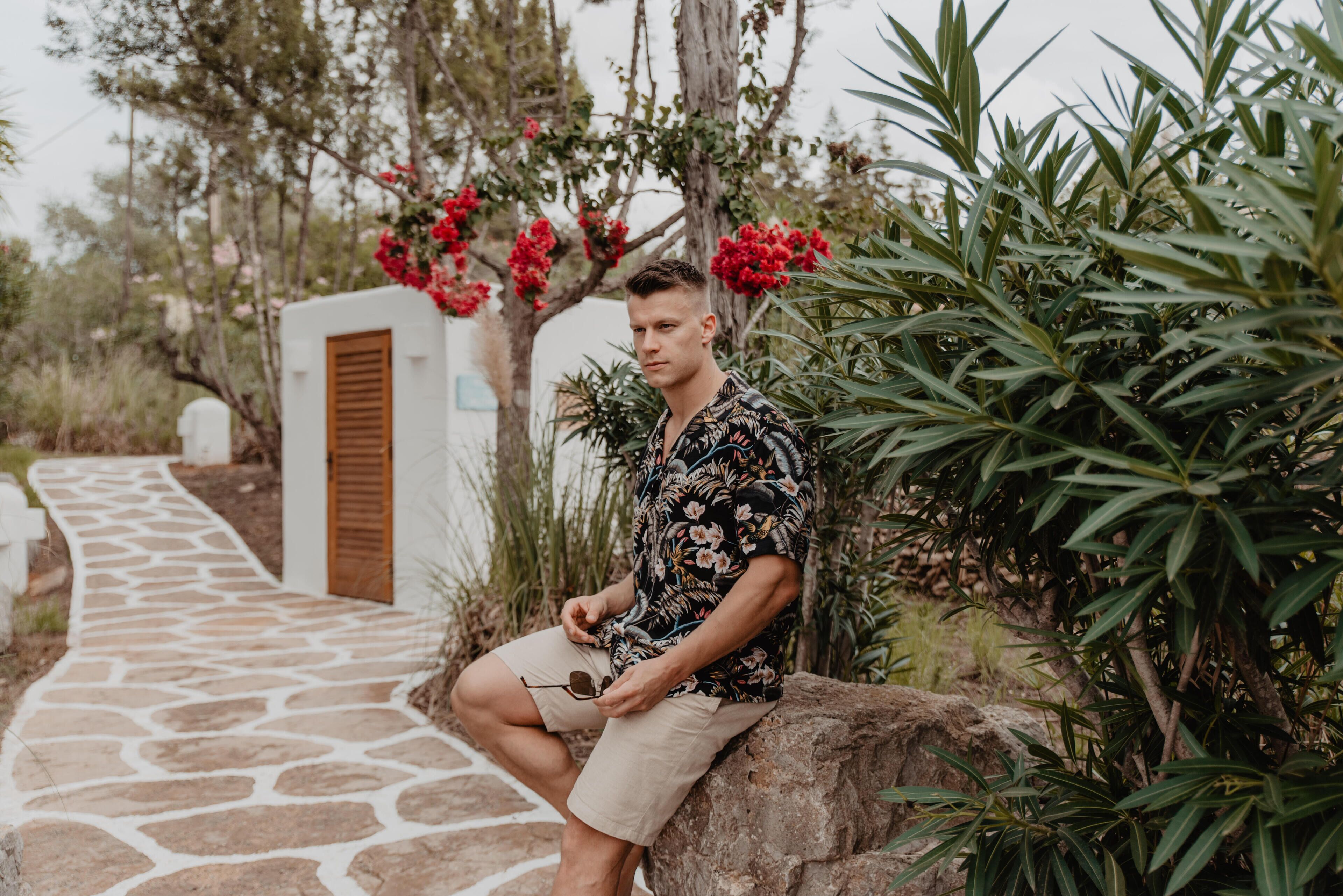 A man sitting on a rock by a stone pathway in a garden with red flowers and green foliage.