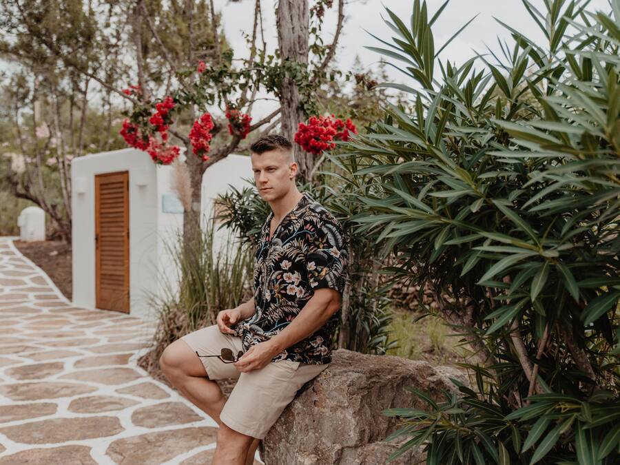 A man sitting on a rock by a stone pathway in a garden with red flowers and green foliage.