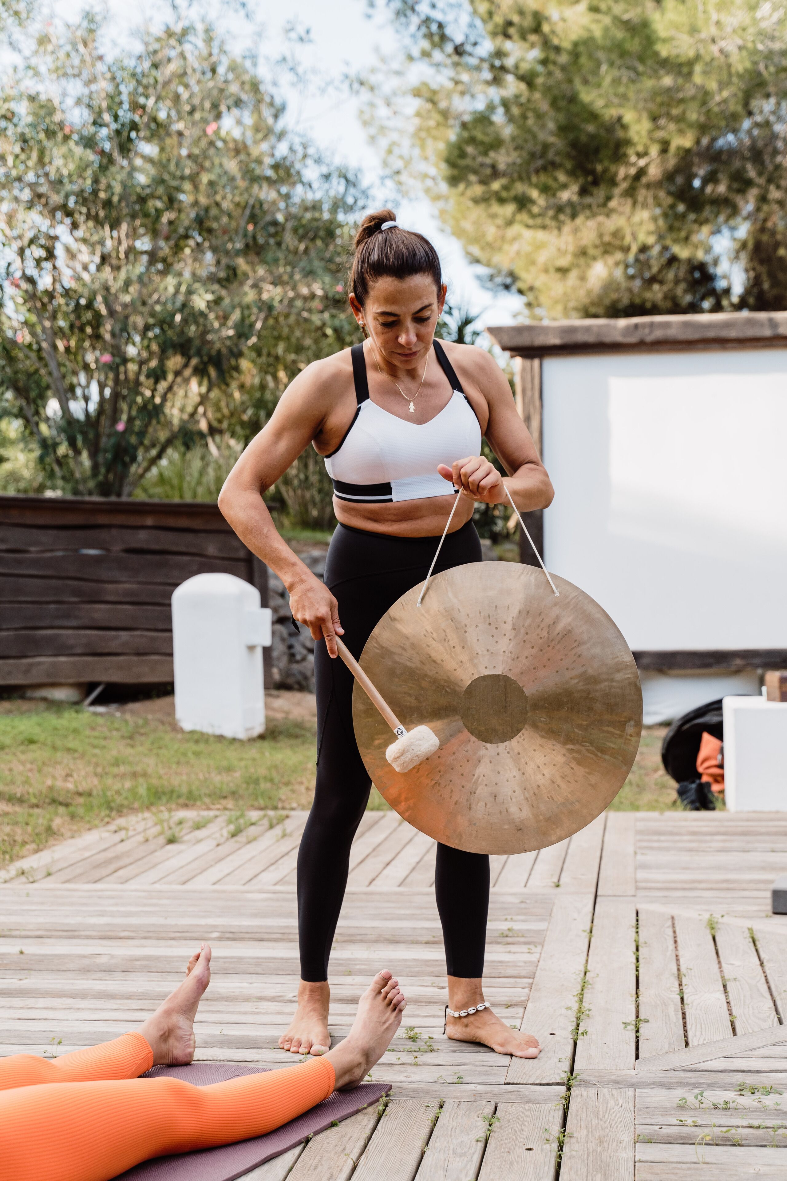 A person standing on a wooden deck holding a large gong, with another person lying on a yoga mat in the foreground.