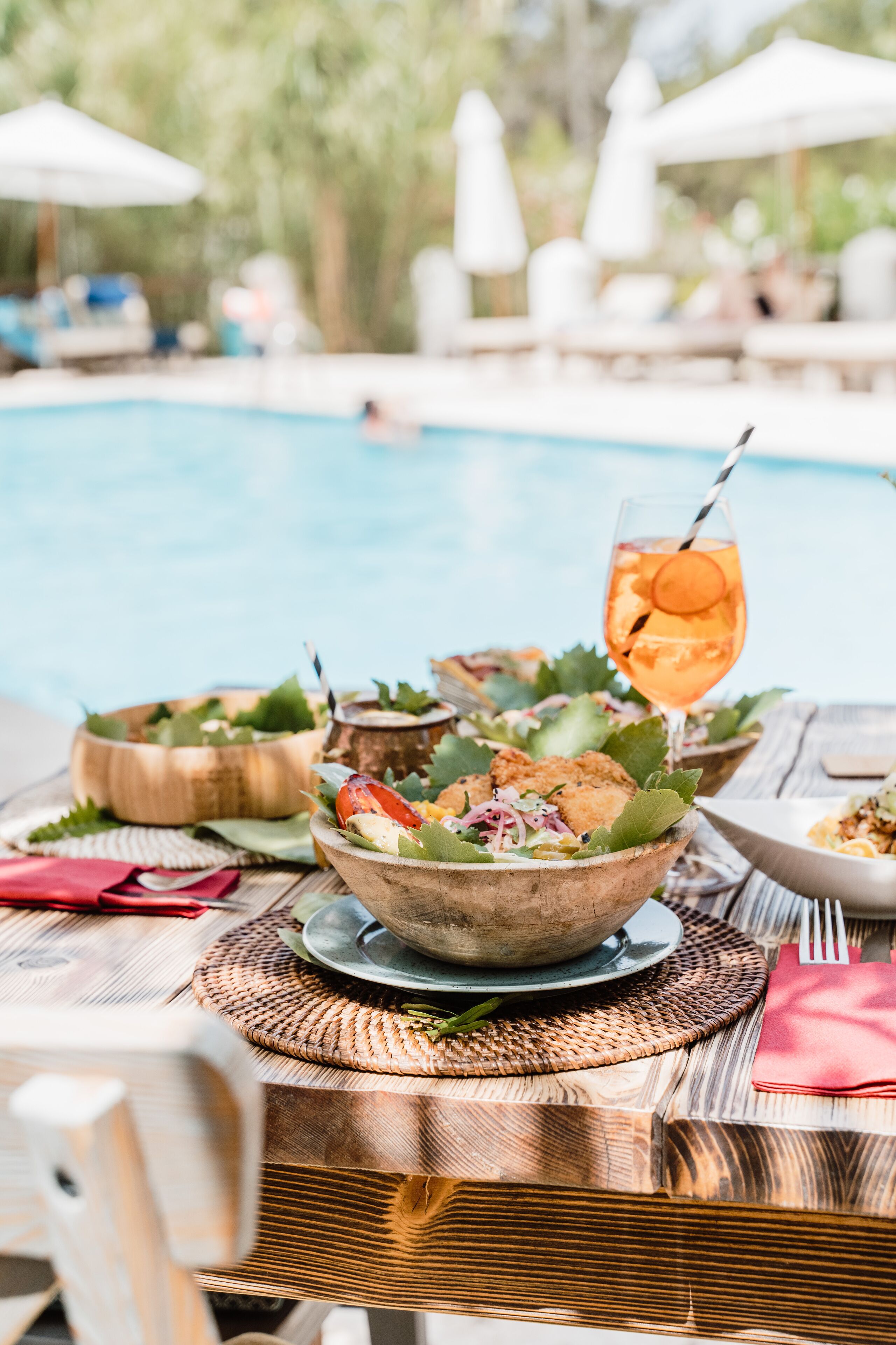 A dining table with a salad, a drink, and a bowl of food, set against a poolside backdrop.