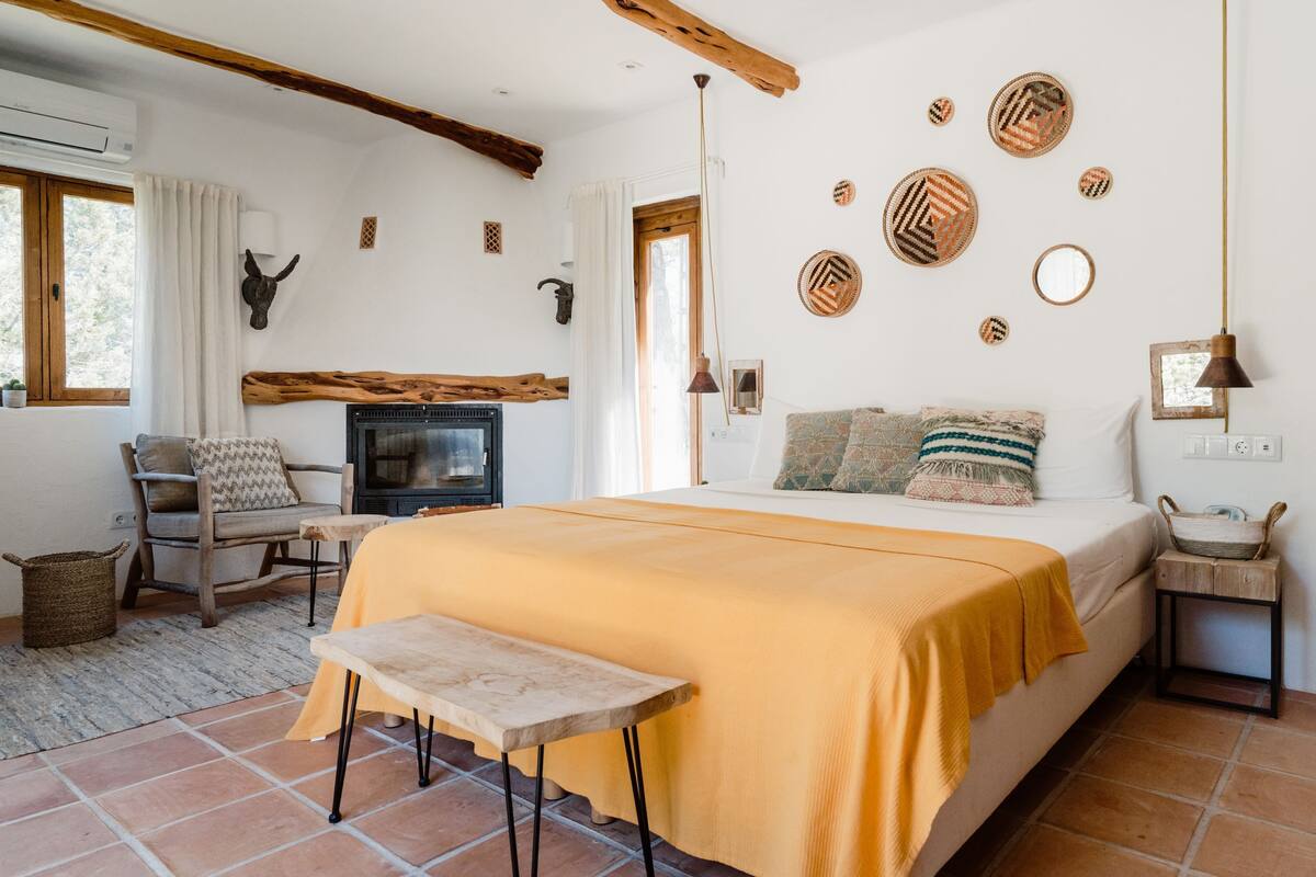 Une chambre confortable avec un lit, un fauteuil à bascule en bois, une petite table, une cheminée et des œuvres d’art décoratives au mur.