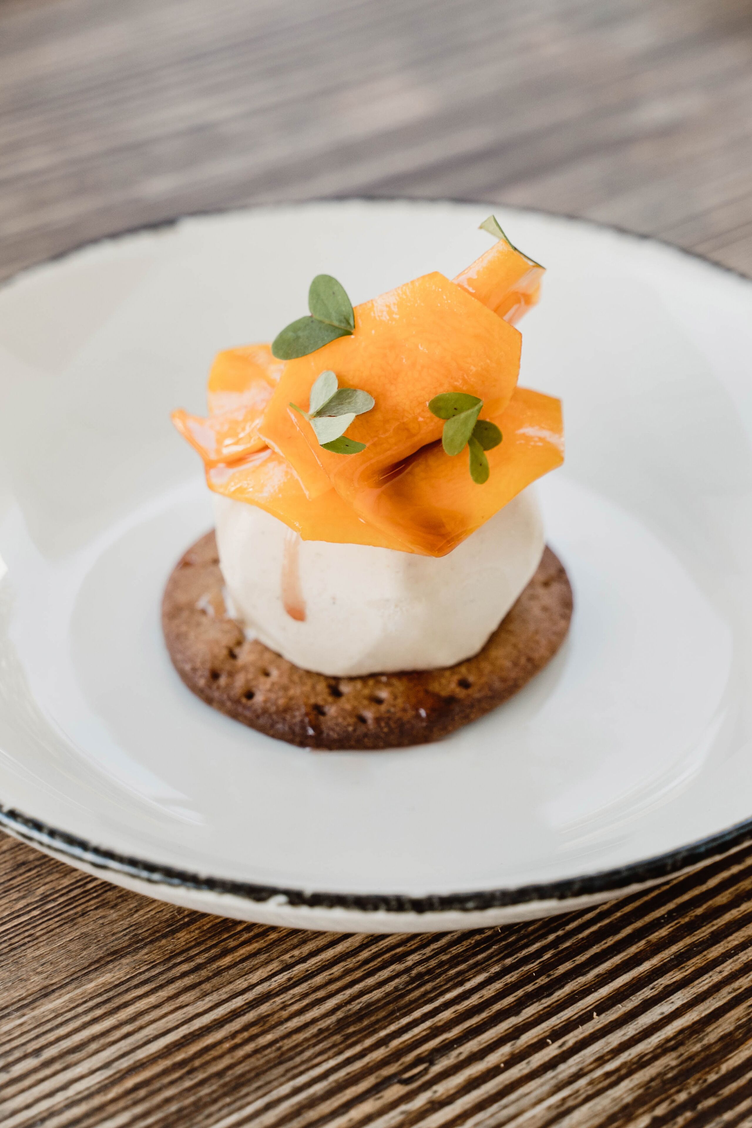 Postre en un plato blanco con borde oscuro, con una porción de helado coronada con rodajas de naranja y hojas verdes.