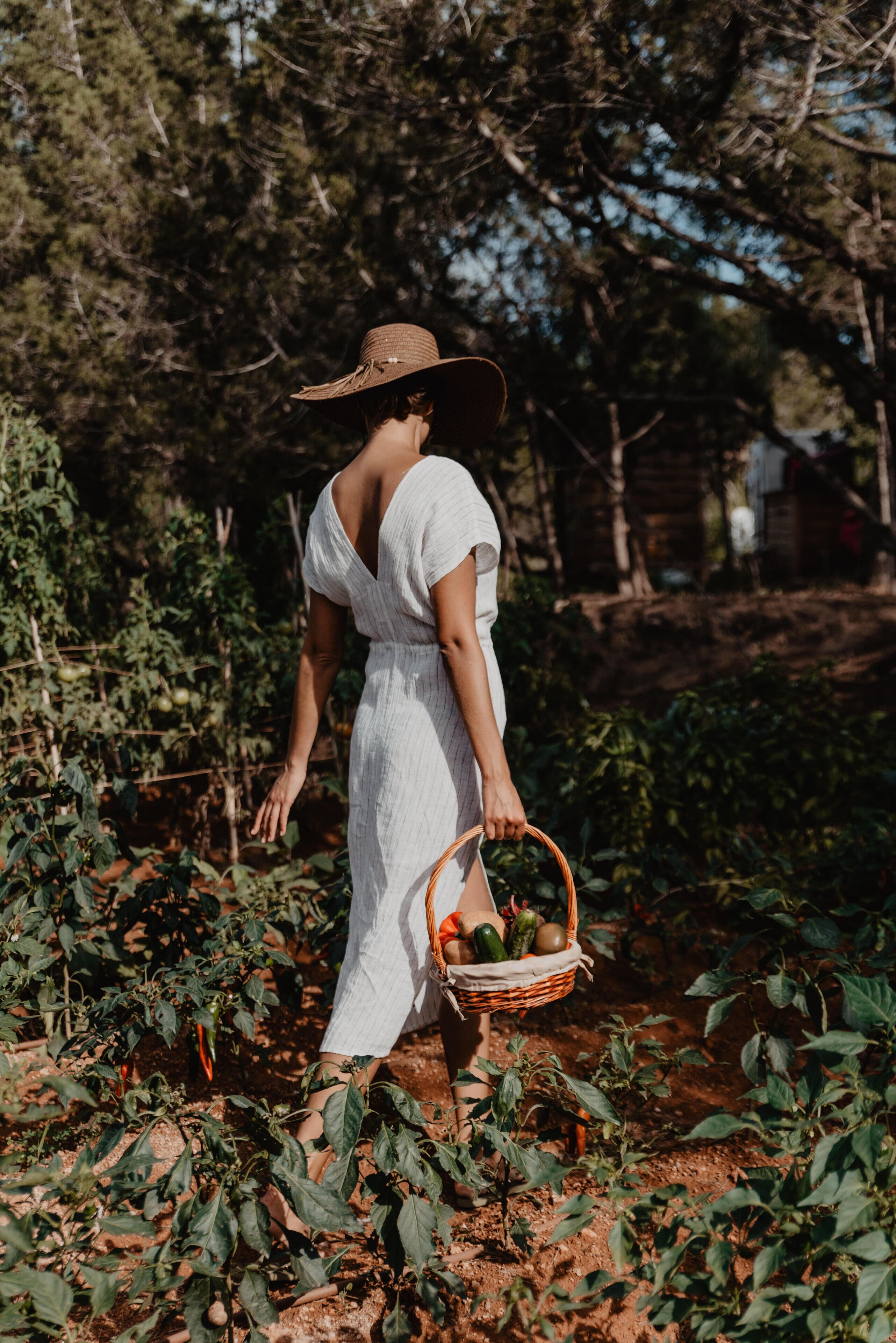 Una persona con vestido blanco y sombrero de ala ancha caminando por un jardín con una cesta de verduras.