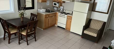 A compact kitchen with wooden cabinets, a stove, a microwave, and a refrigerator. There is a dining table with chairs and a small seating area with a cushion.