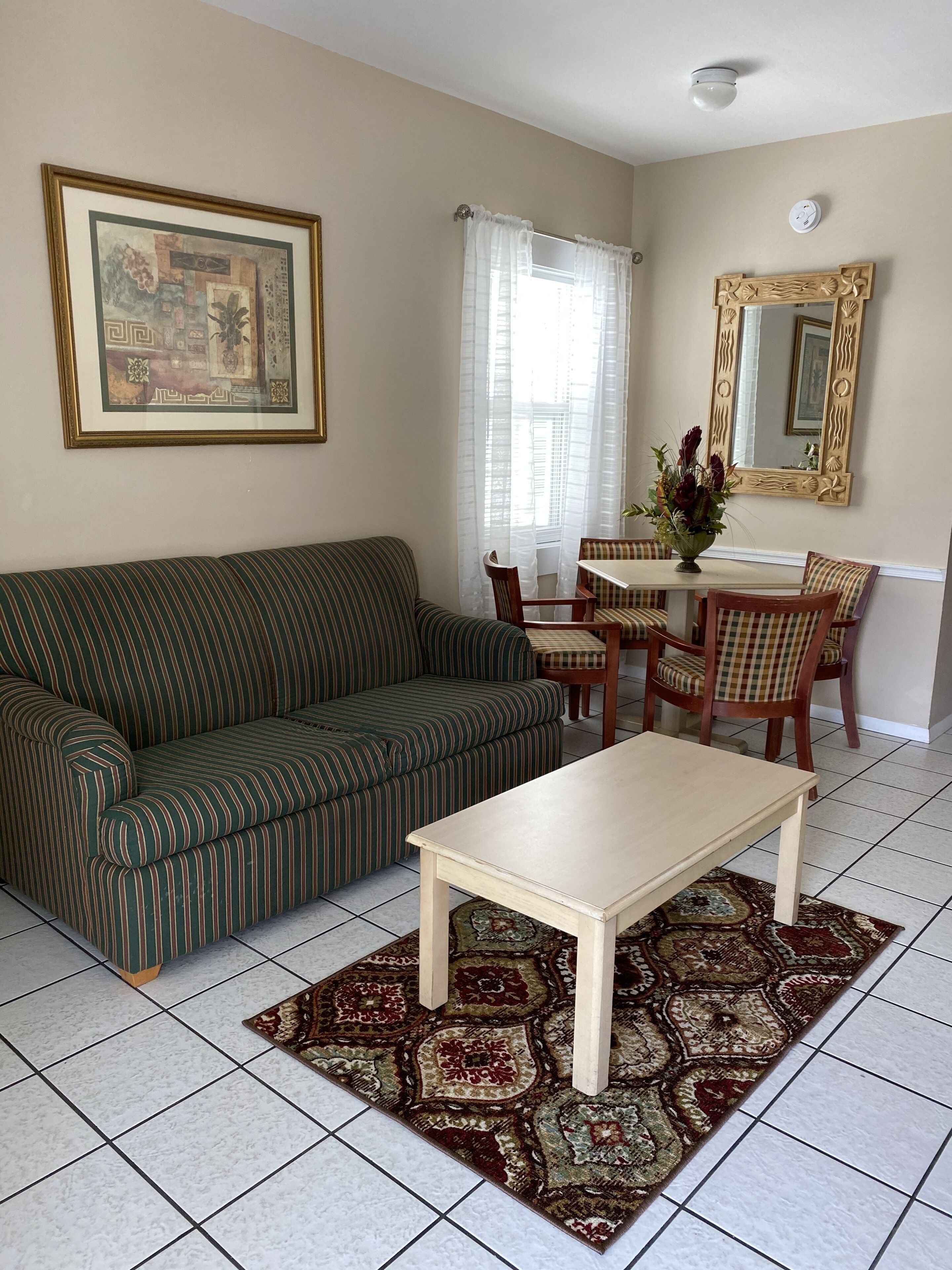 A living room with a striped sofa, a white coffee table, a dining table with chairs, a large mirror, and a patterned rug.
