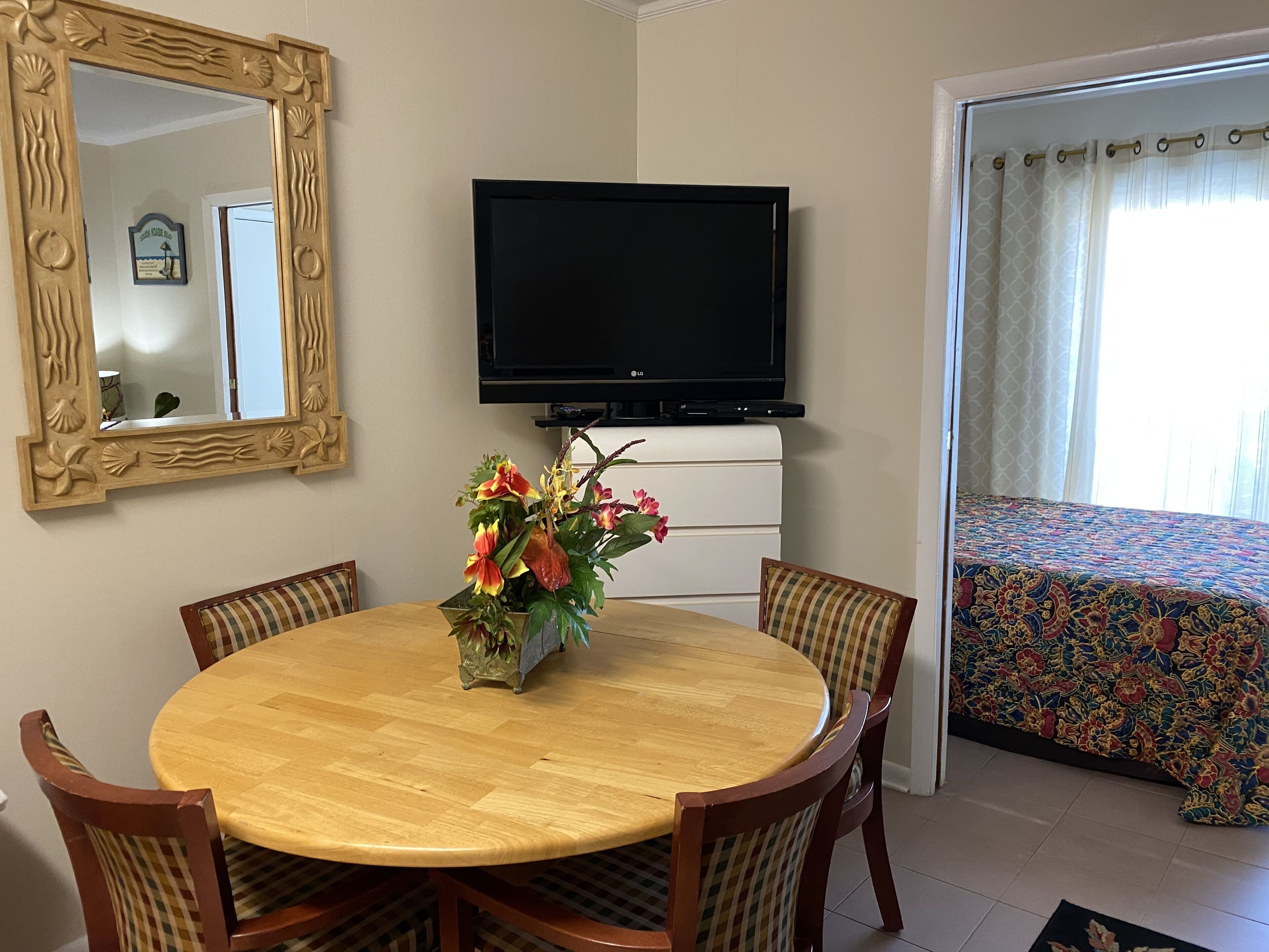 A room with a round wooden dining table, four chairs, a television on the wall, a floral centerpiece, and a bed with a patterned bedspread.