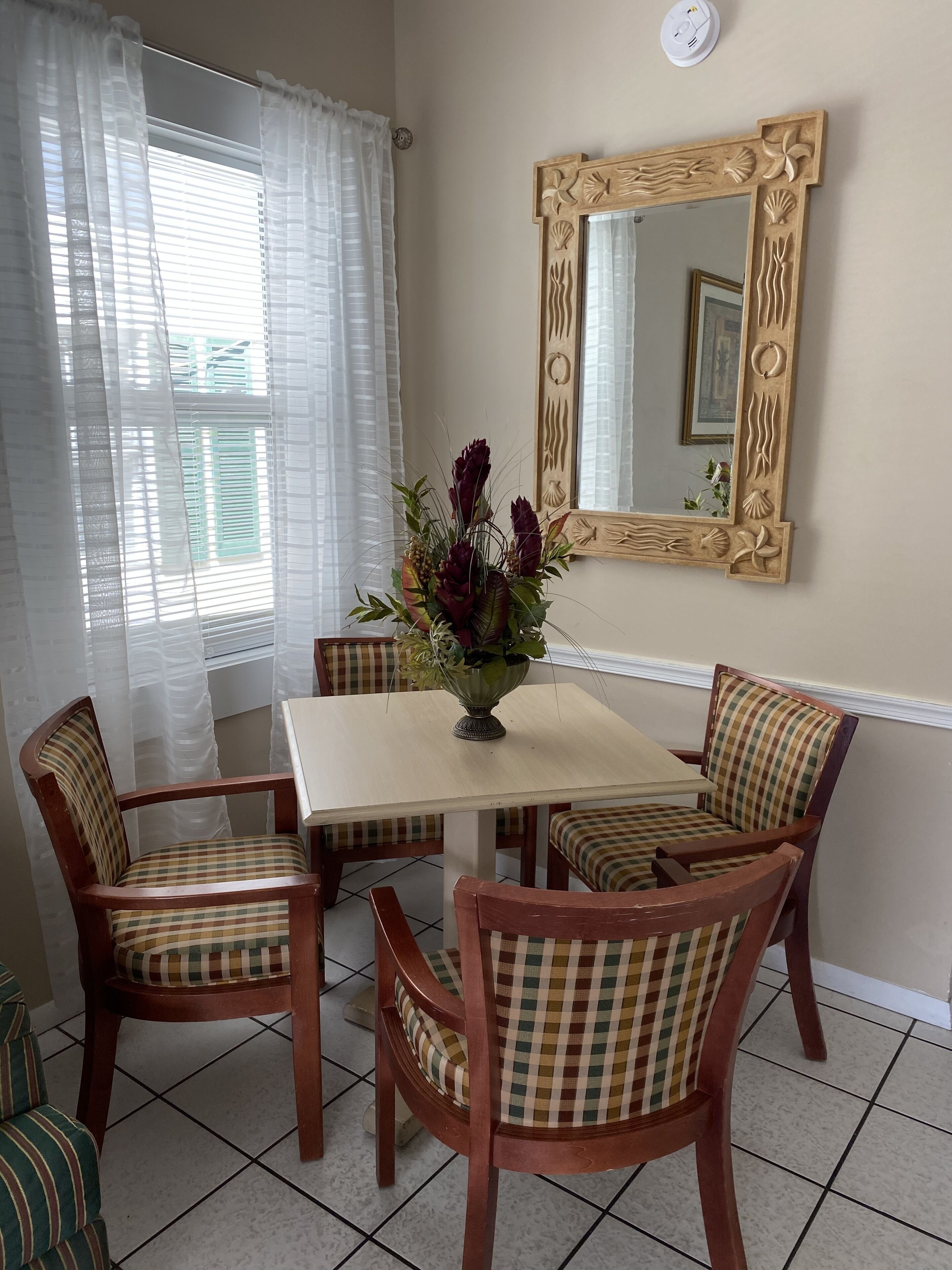 Un comedor con una mesa y cuatro sillas, un jarrón con flores, un espejo con marco decorativo y una ventana con cortinas traslúcidas.