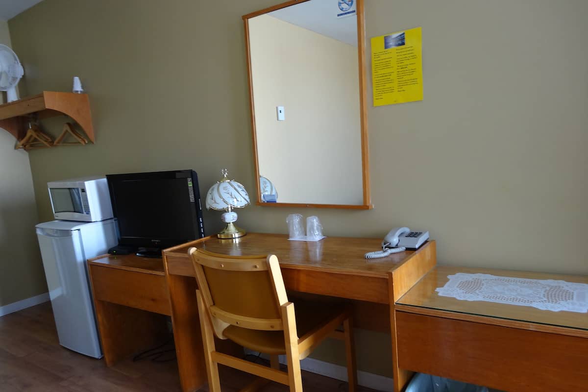 A compact hotel room with a wooden desk, a computer, a small fridge, a chair, and a mirror.