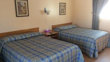 A room with two beds, each with a patterned blue and gold bedspread, a nightstand with a tissue box, and a wall-mounted lamp.