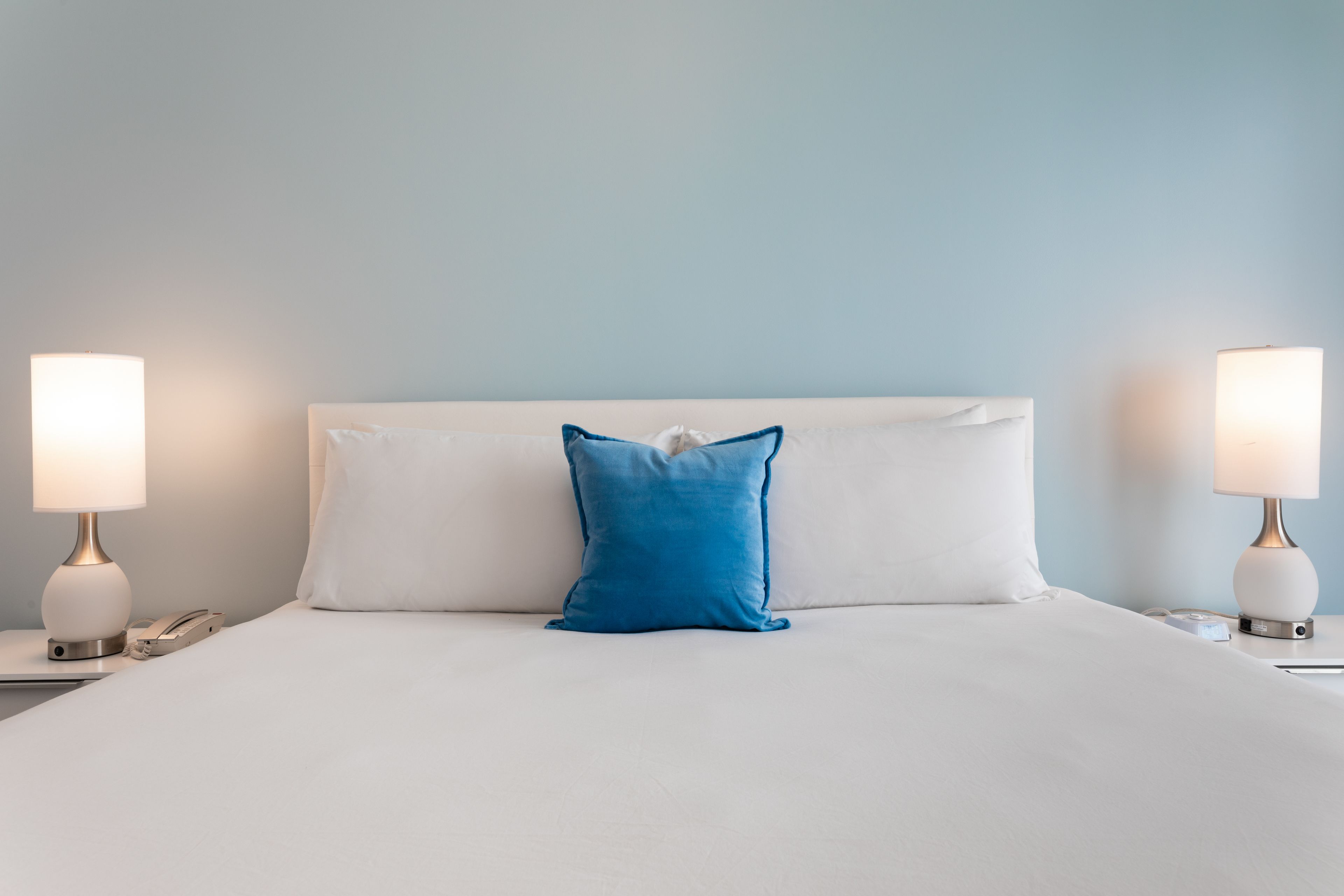 Un lit bien fait, avec du linge de lit blanc et un seul oreiller bleu, flanqué de deux tables de chevet sur lesquelles sont posées des lampes et un téléphone.