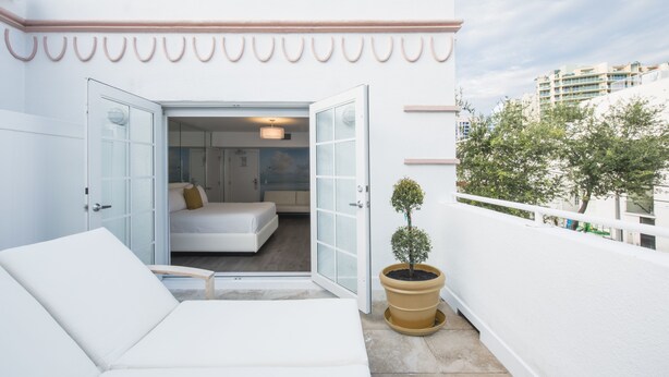 Un balcon moderne avec un canapé blanc, une petite plante en pot et un aperçu d’une chambre à coucher avec un lit et une table de chevet.