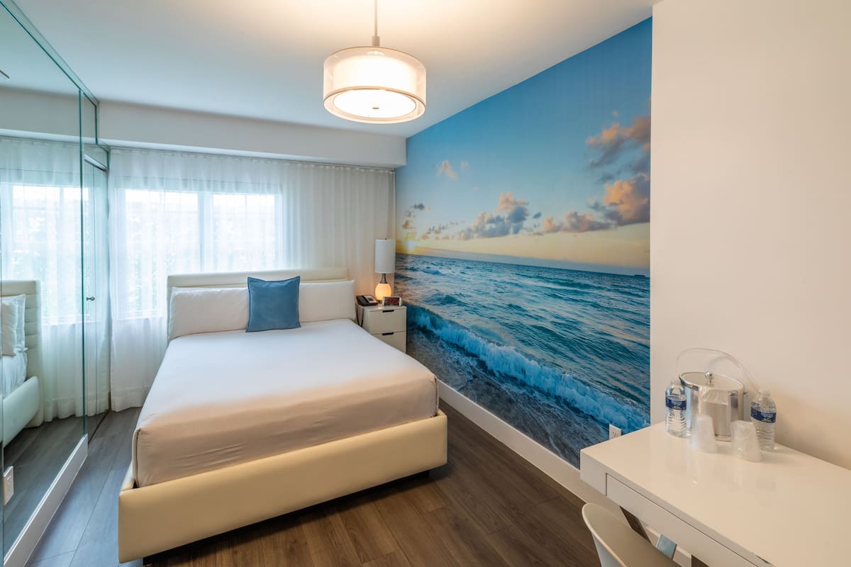 A hotel room with a bed, a nightstand, a desk, and a large ocean mural on the wall.