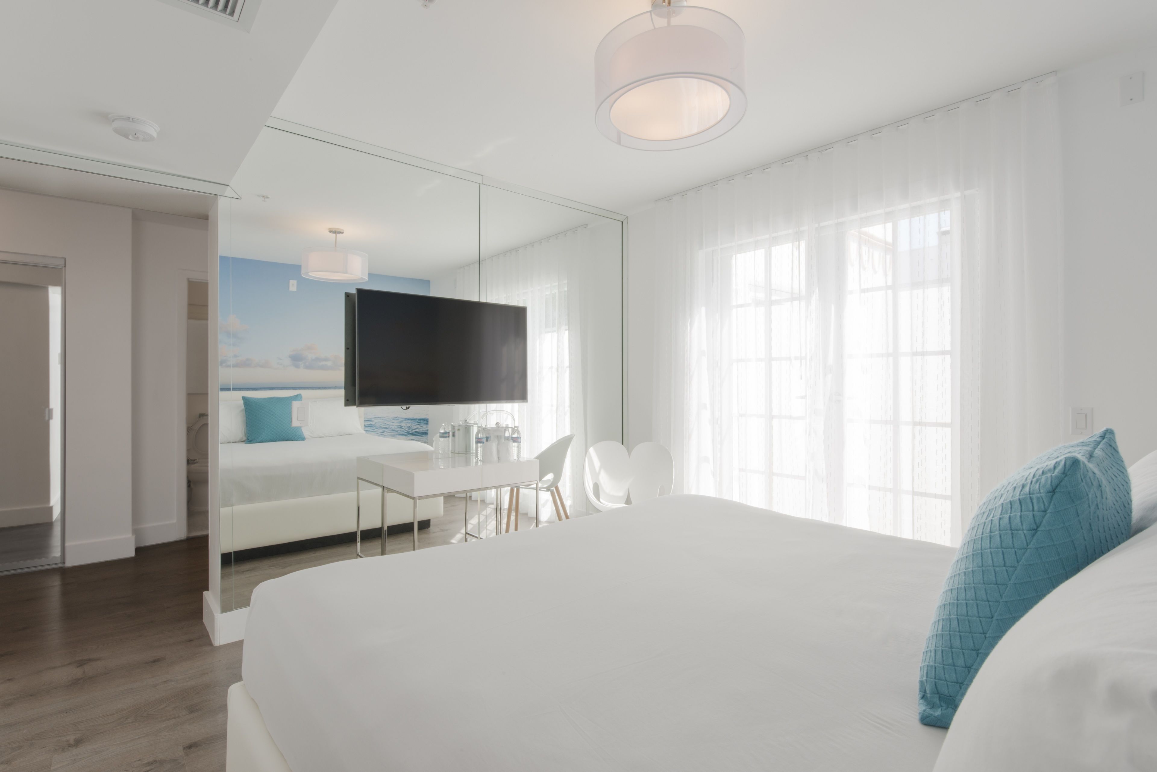 A modern hotel room with a large bed, a desk, a chair, a flat-screen TV, and a large window with sheer curtains.