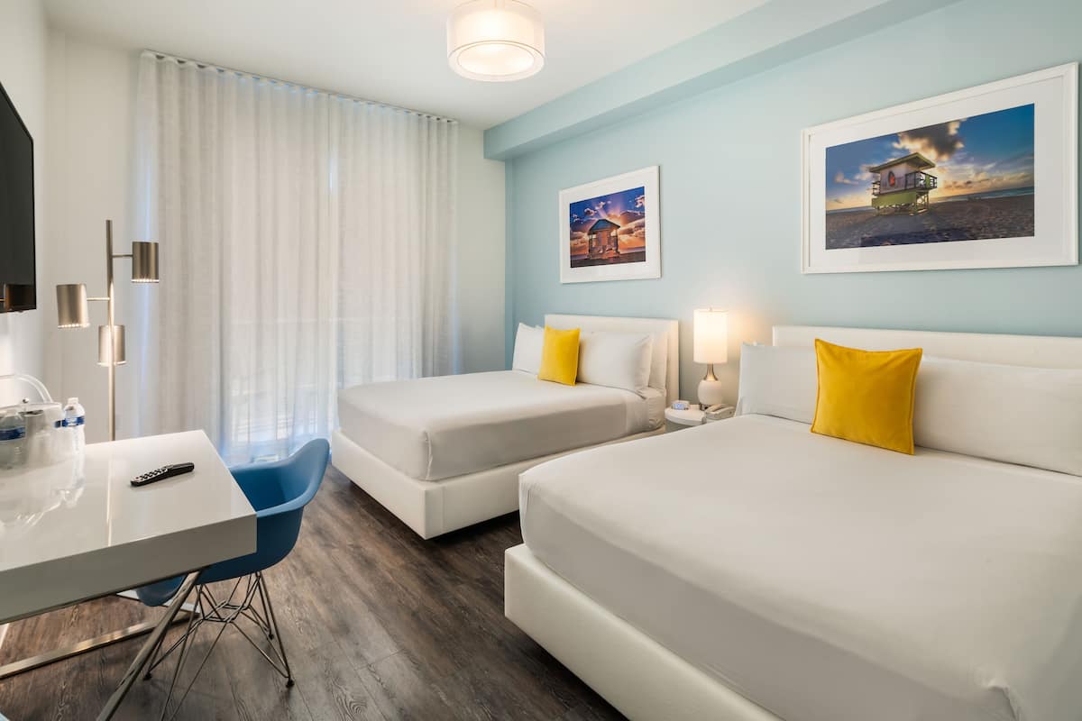A hotel room with two beds, a desk, and a chair. There are two paintings on the wall and a window with curtains.