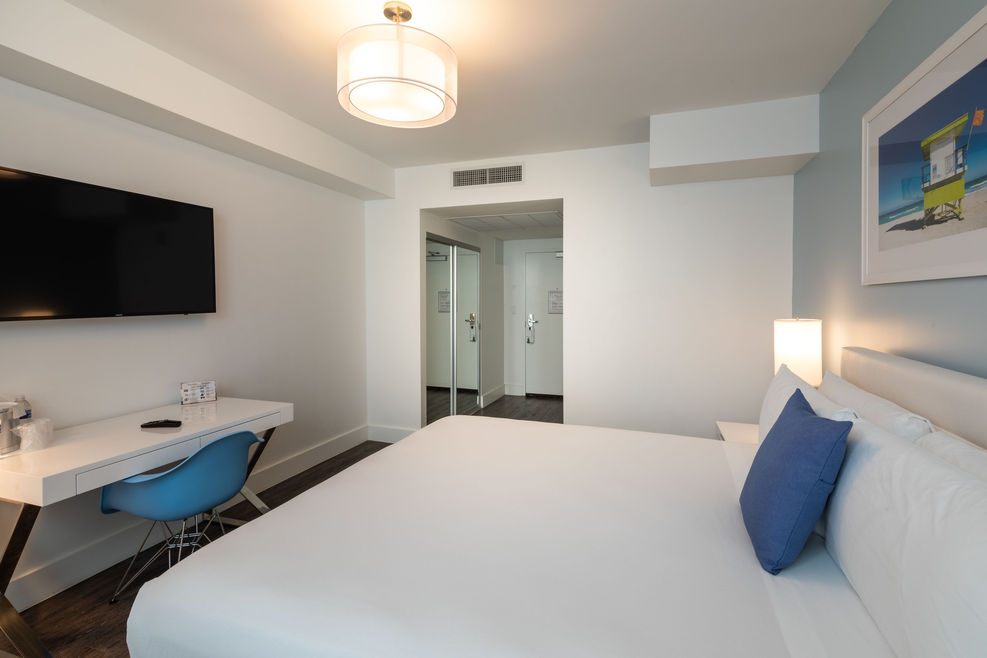 Ein modernes Hotelzimmer mit einem großen Bett, einem Schreibtisch, einem blauen Sessel, einem Flachbildfernseher und einem gerahmten Bild an der Wand.