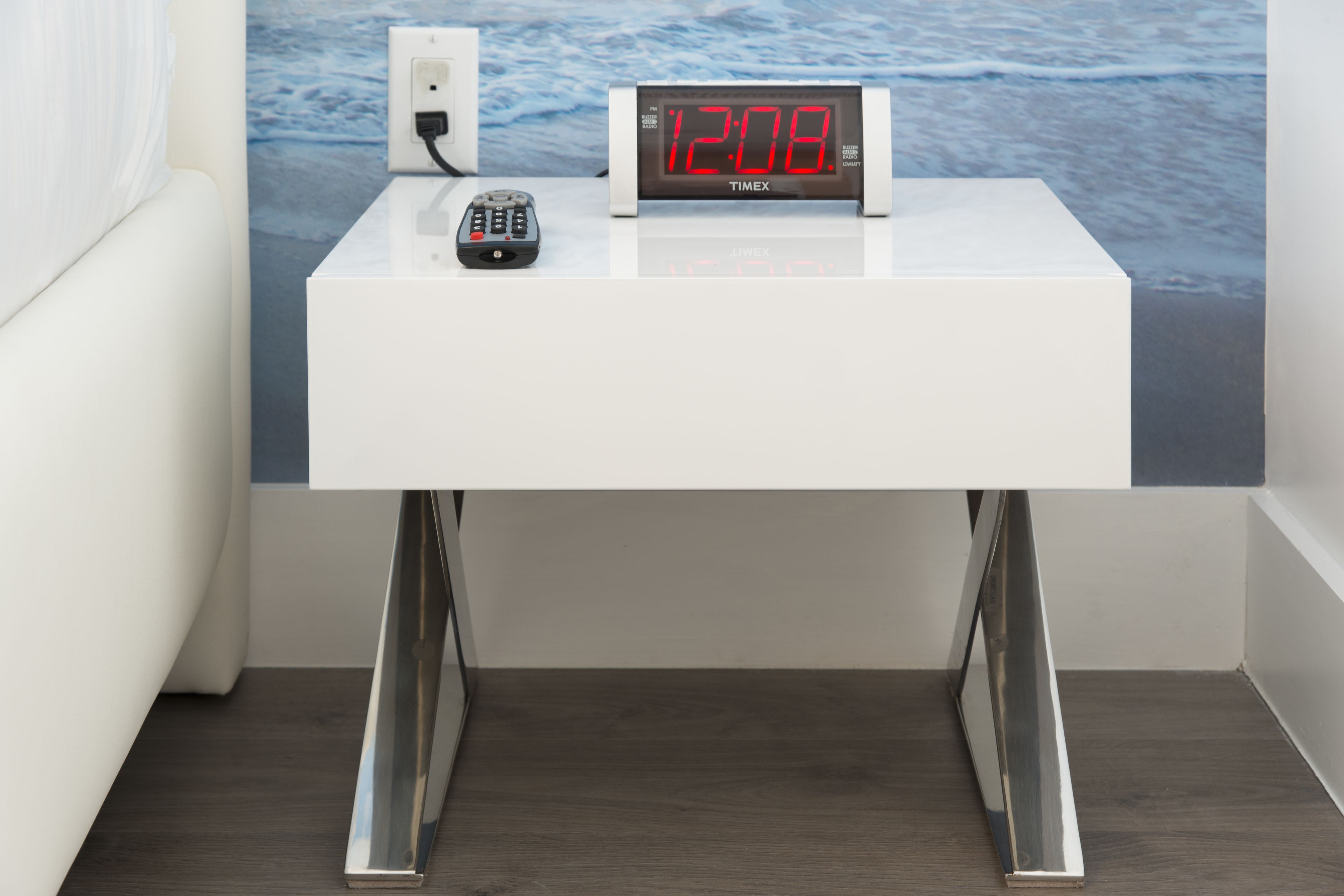 A white bedside table with a digital clock displaying 12:08, a remote control, and a wall outlet.
