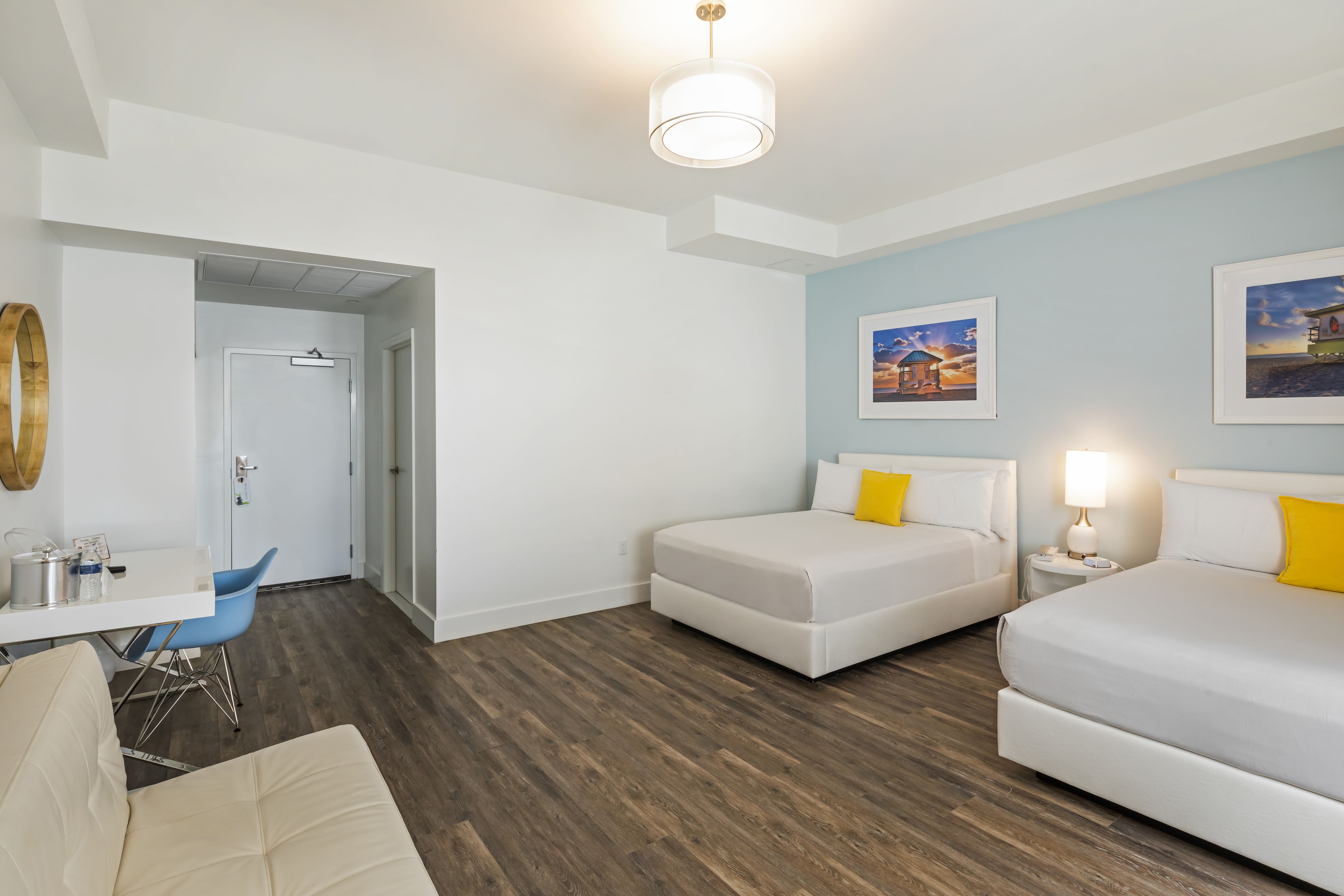 A modern hotel room with a wooden floor, a white bed, a small desk, and a chair. There is a door leading to another room and a wall with two framed pictures.