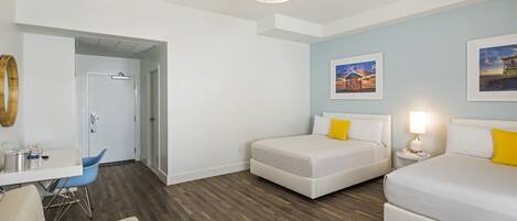 A modern hotel room with a wooden floor, a white bed, a small desk, and a chair. There is a door leading to another room and a wall with two framed pictures.
