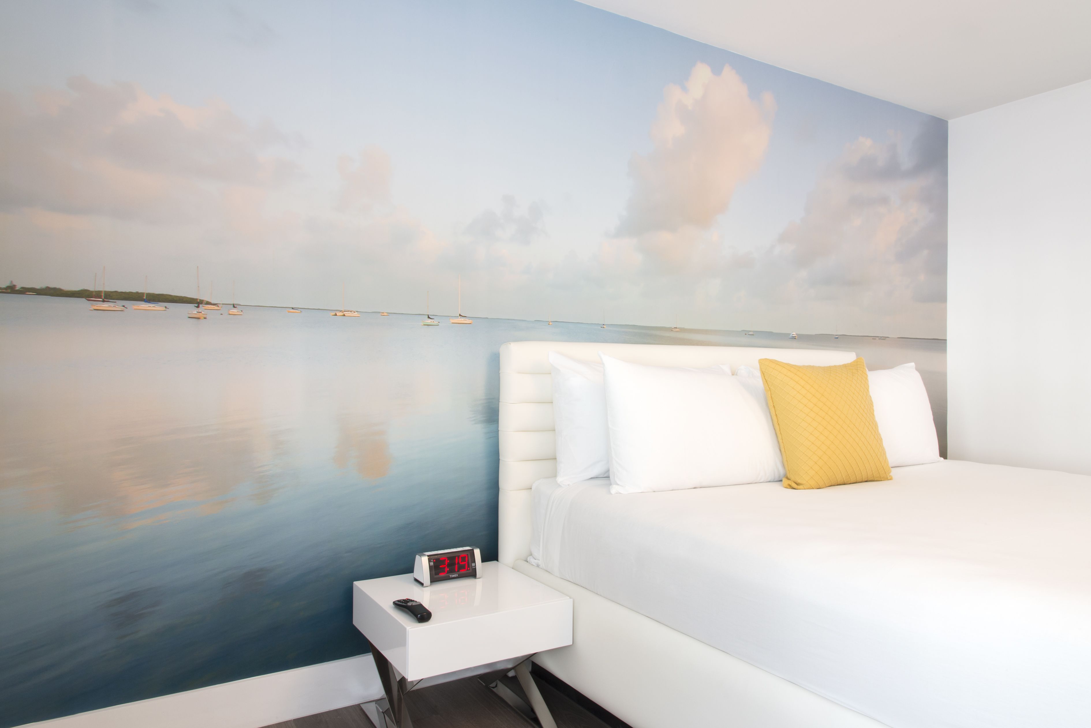 Une chambre à coucher ornée d’une fresque représentant une mer calme et des bateaux, avec un lit blanc et un oreiller jaune, ainsi qu’une table de chevet sur laquelle se trouvent une horloge et une télécommande.